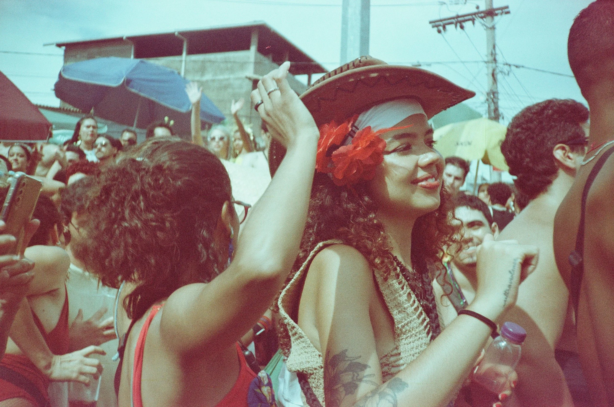 Foliã de chapéu e flor vermelha sorrindo em meio à multidão do bloco Como te Lhama no Carnaval 2026, em filme Solaris 100 da Festival de filmes vencidos