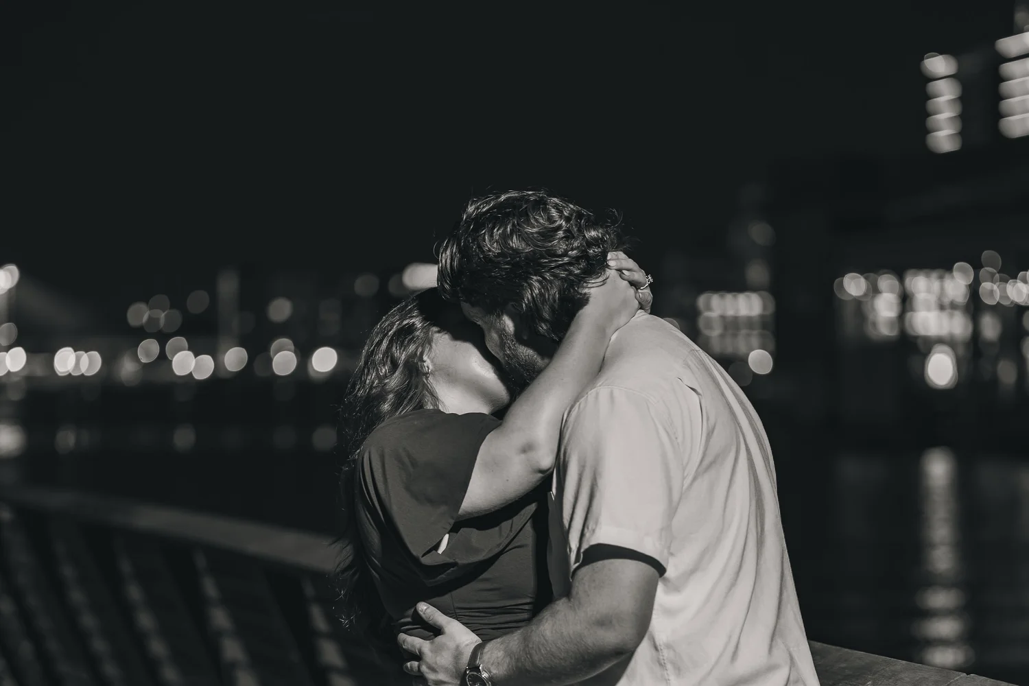 A newly engaged couple sharing a kiss at night with city lights behind them