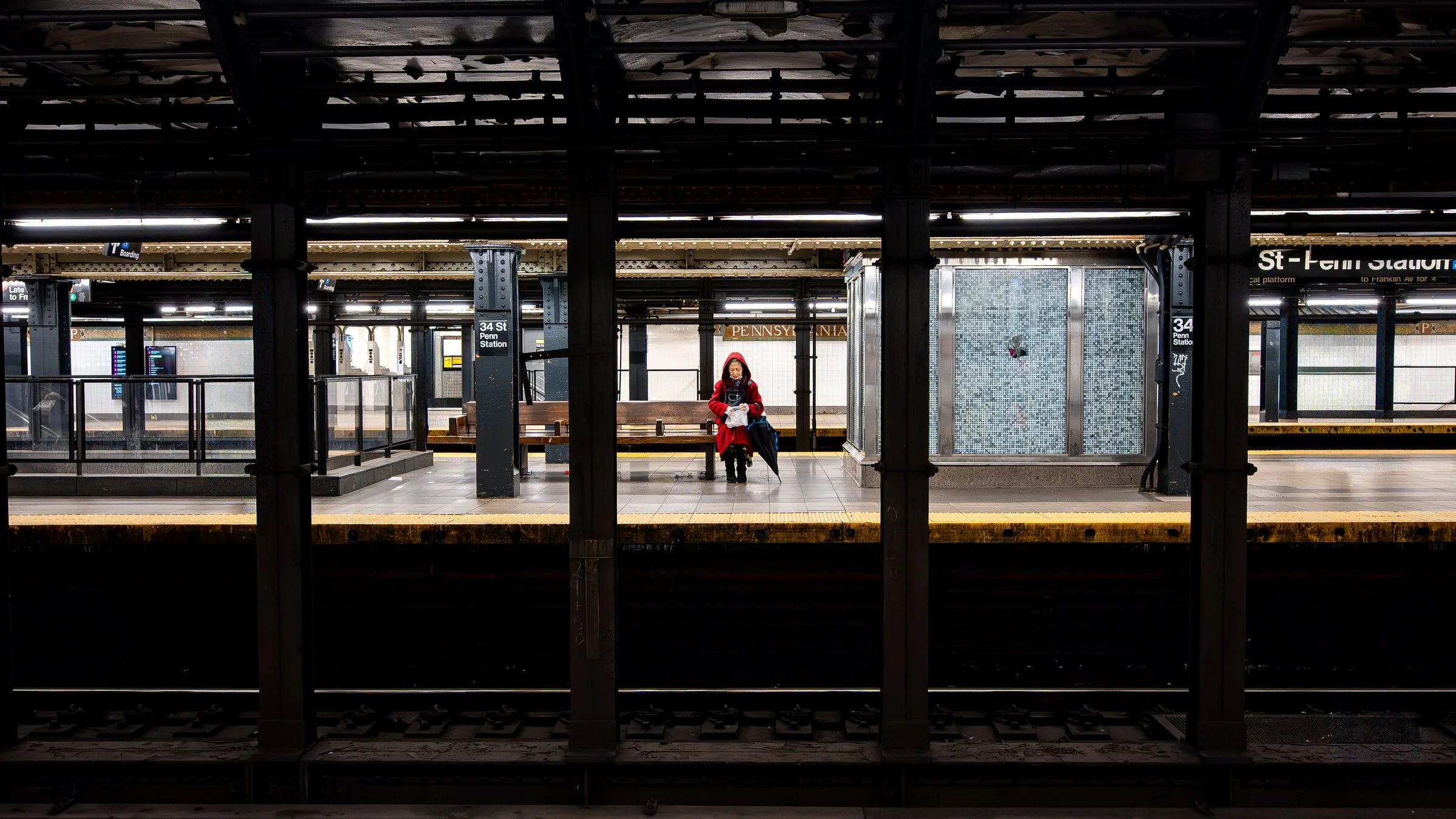 A woman sitting on a bench at Pennsylvania train station, wearing a red coat and holding a white bag, with an empty platform behind her and the station sign above.