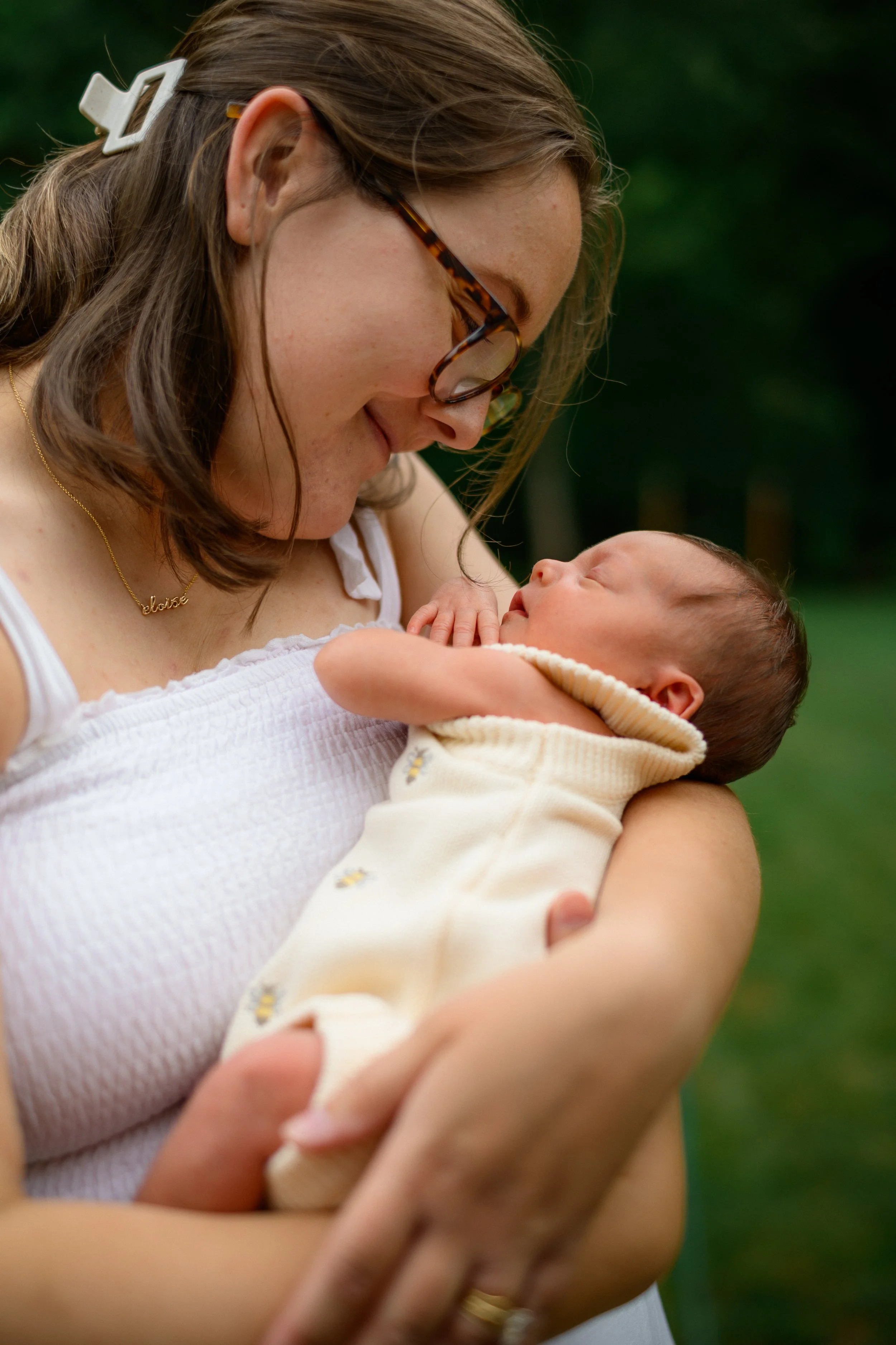 A woman with brown hair, glasses, and a gold necklace holding a newborn baby outside in a green park.