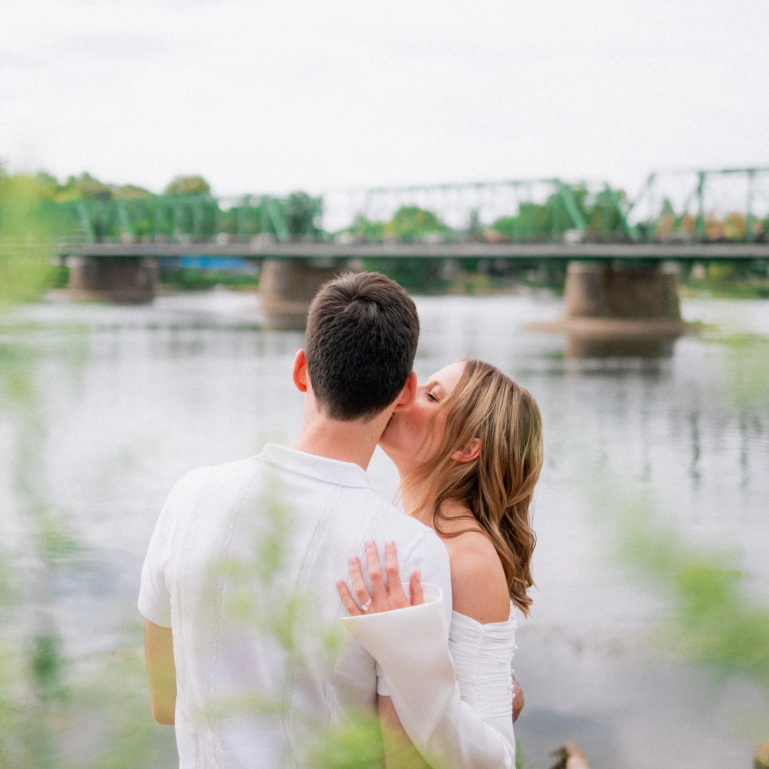 A couple sharing a kiss by a river with a green bridge behind them