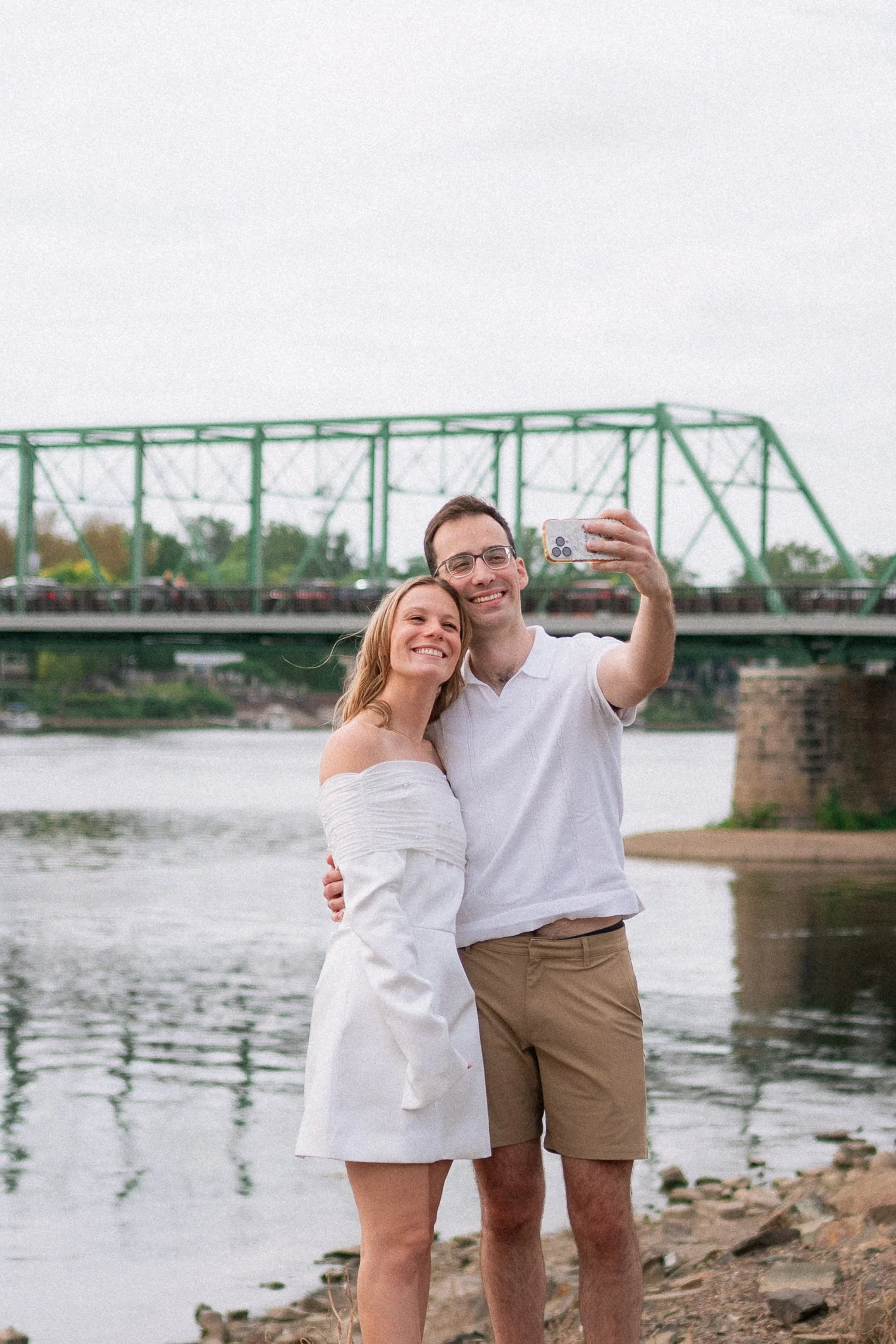 A couple taking a selfie on a rocky riverbank with a bridge behind them