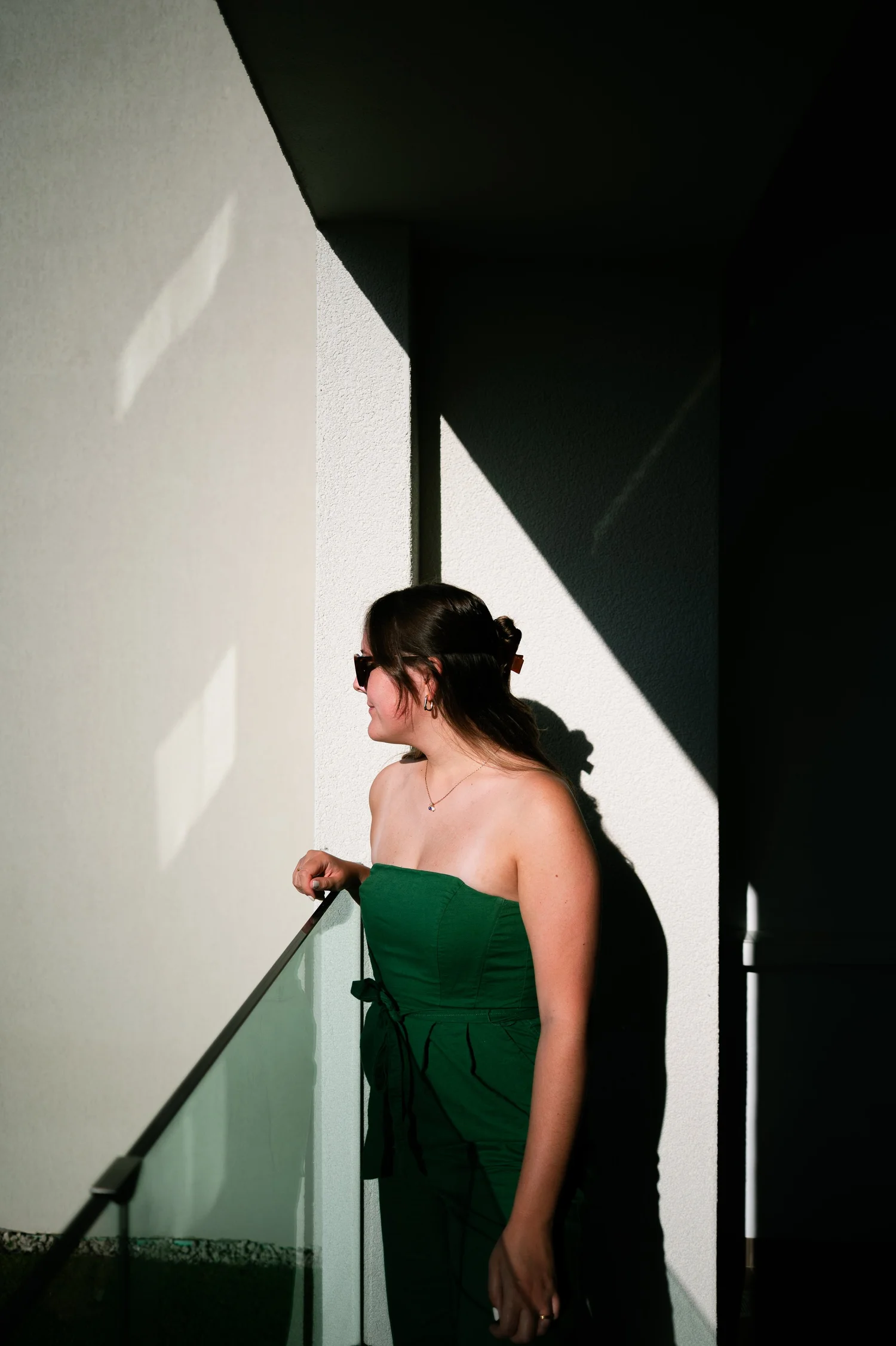 A woman in a green strapless dress and sunglasses against a sunlit white wall