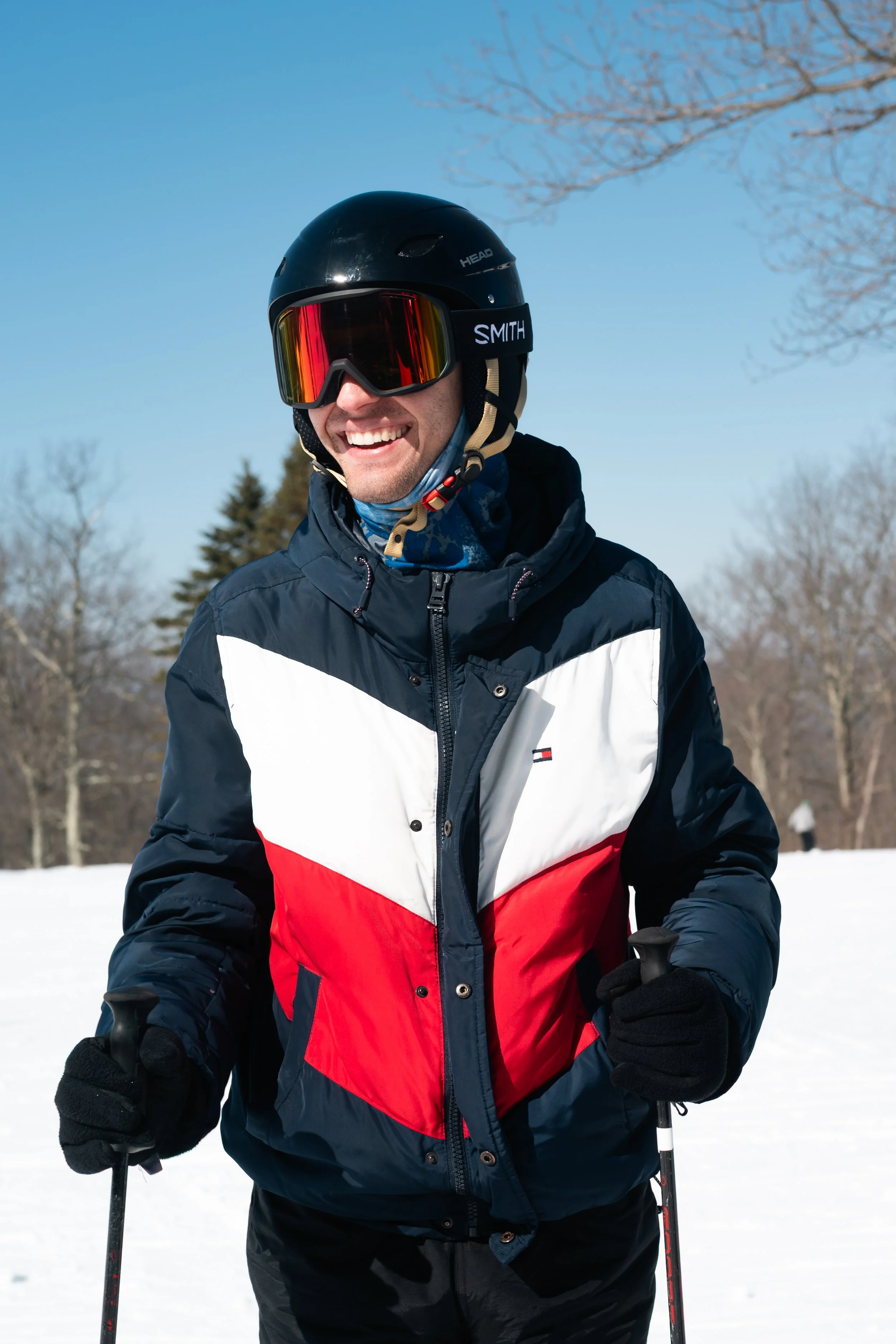 A man wearing a black helmet, ski goggles, gloves, and a color-blocked winter jacket smiling outdoors in a snowy landscape.
