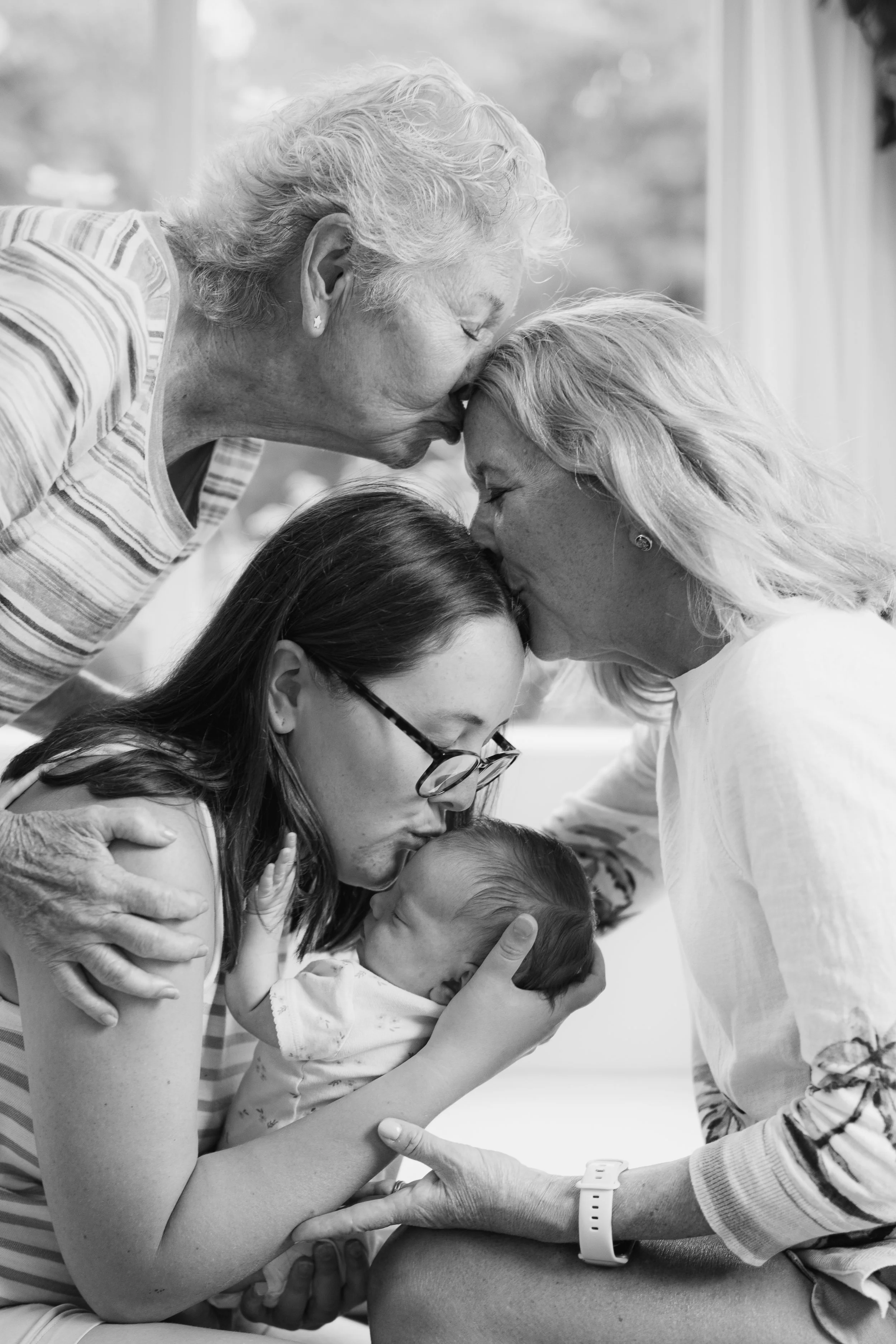 An elderly woman and elderly man, both with white hair, share a kiss on the forehead while holding a young child, a girl with glasses, suggestive of a family moment.