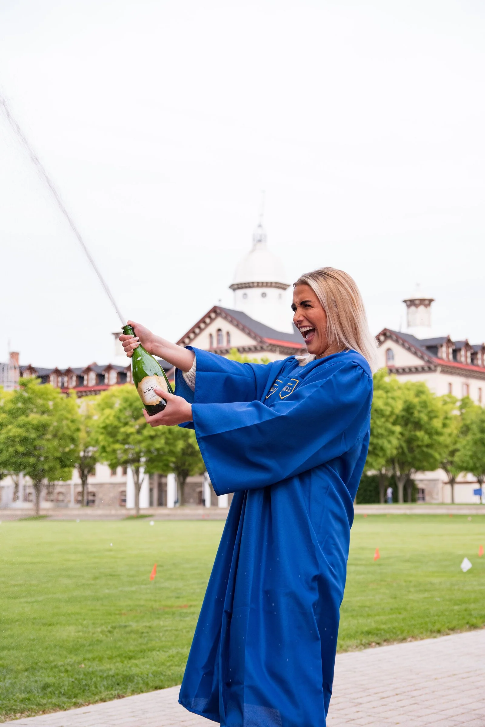 A woman in a blue graduation gown holding a champagne bottle, celebrating outdoors on a cloudy day.