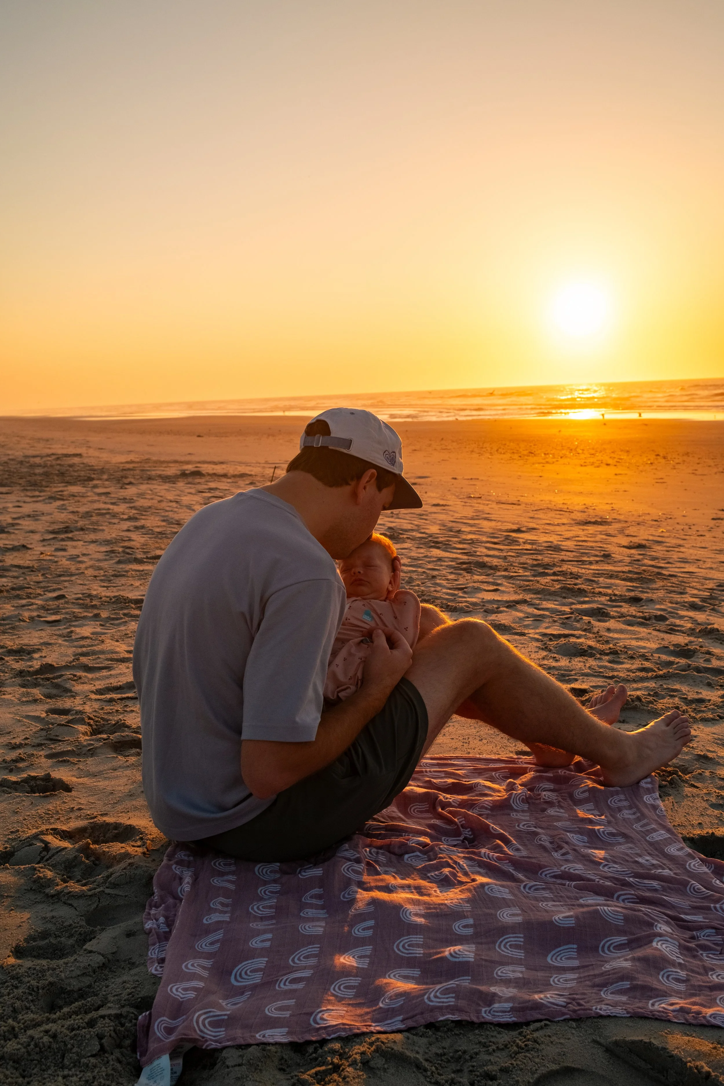 A man holding a baby on a beach at sunset with the ocean in the background.