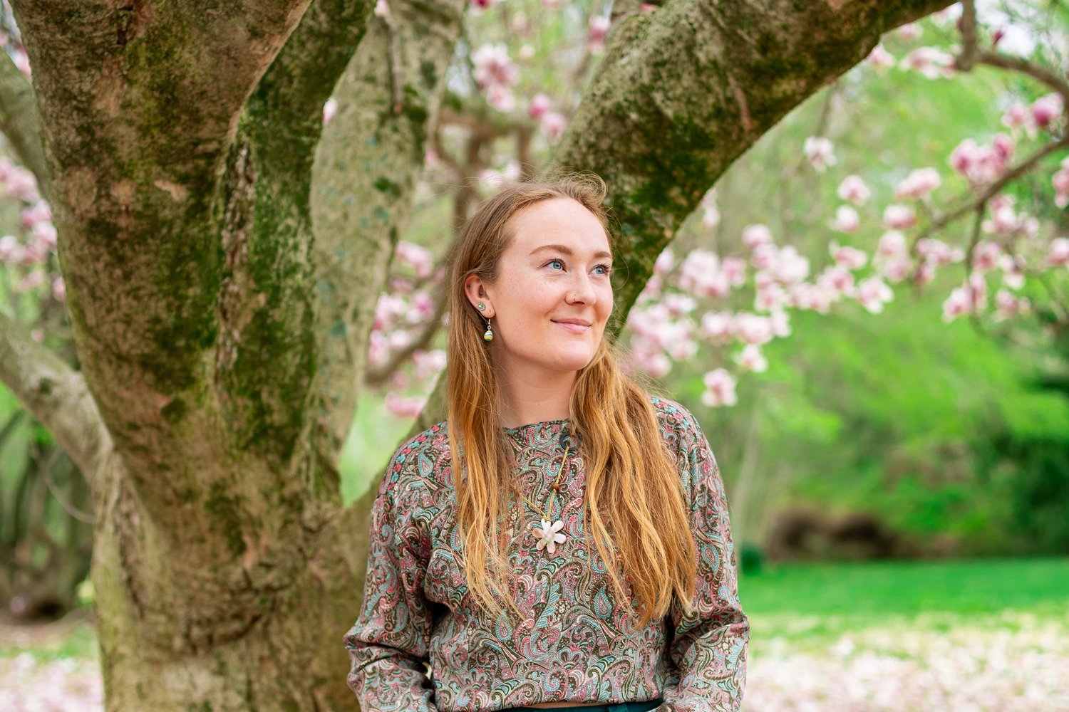 A woman with long red hair smiling beneath a tree in pink blossom