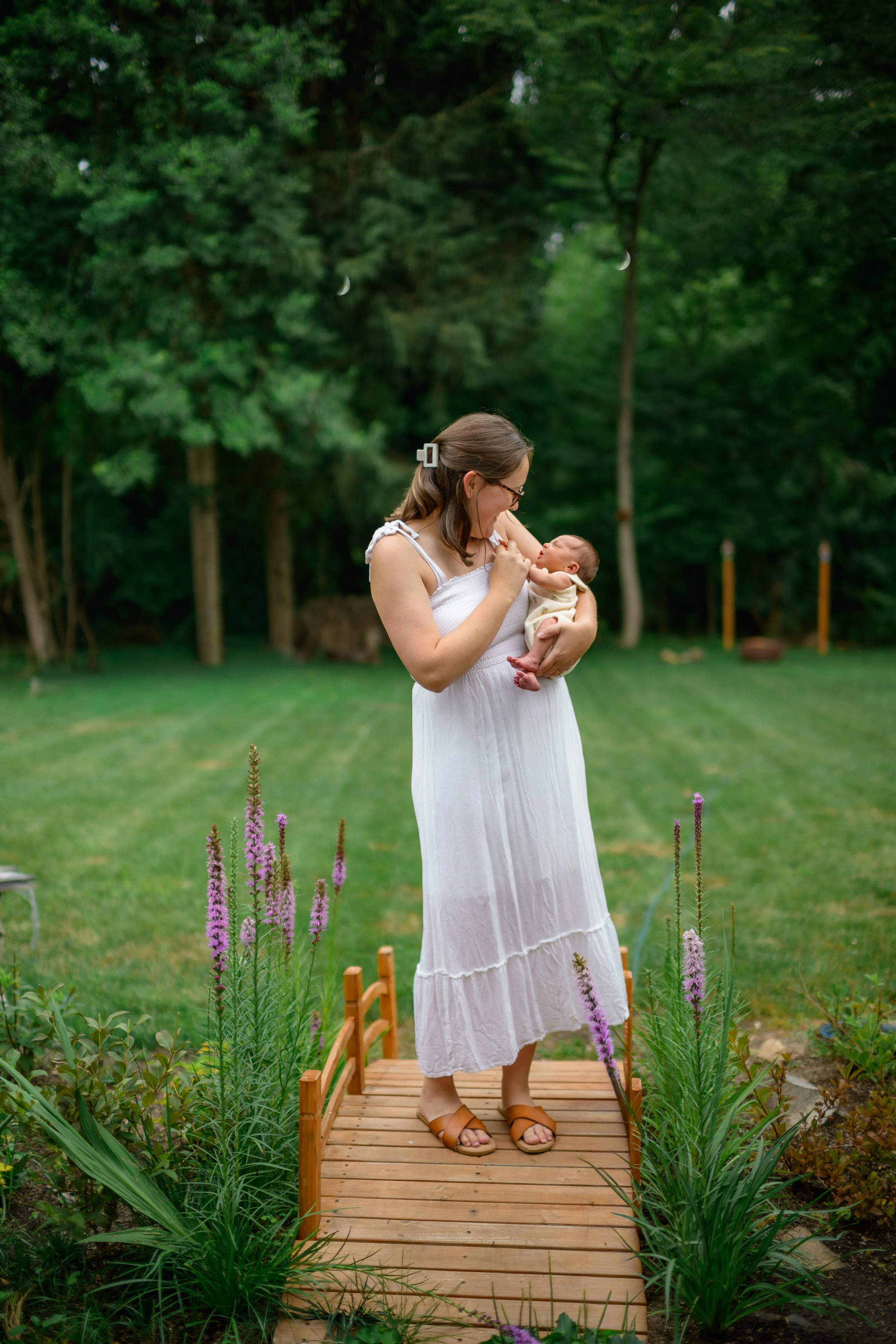A woman in a long white dress holding a baby standing on a small wooden bridge in a garden with green grass, trees, and purple flowers.
