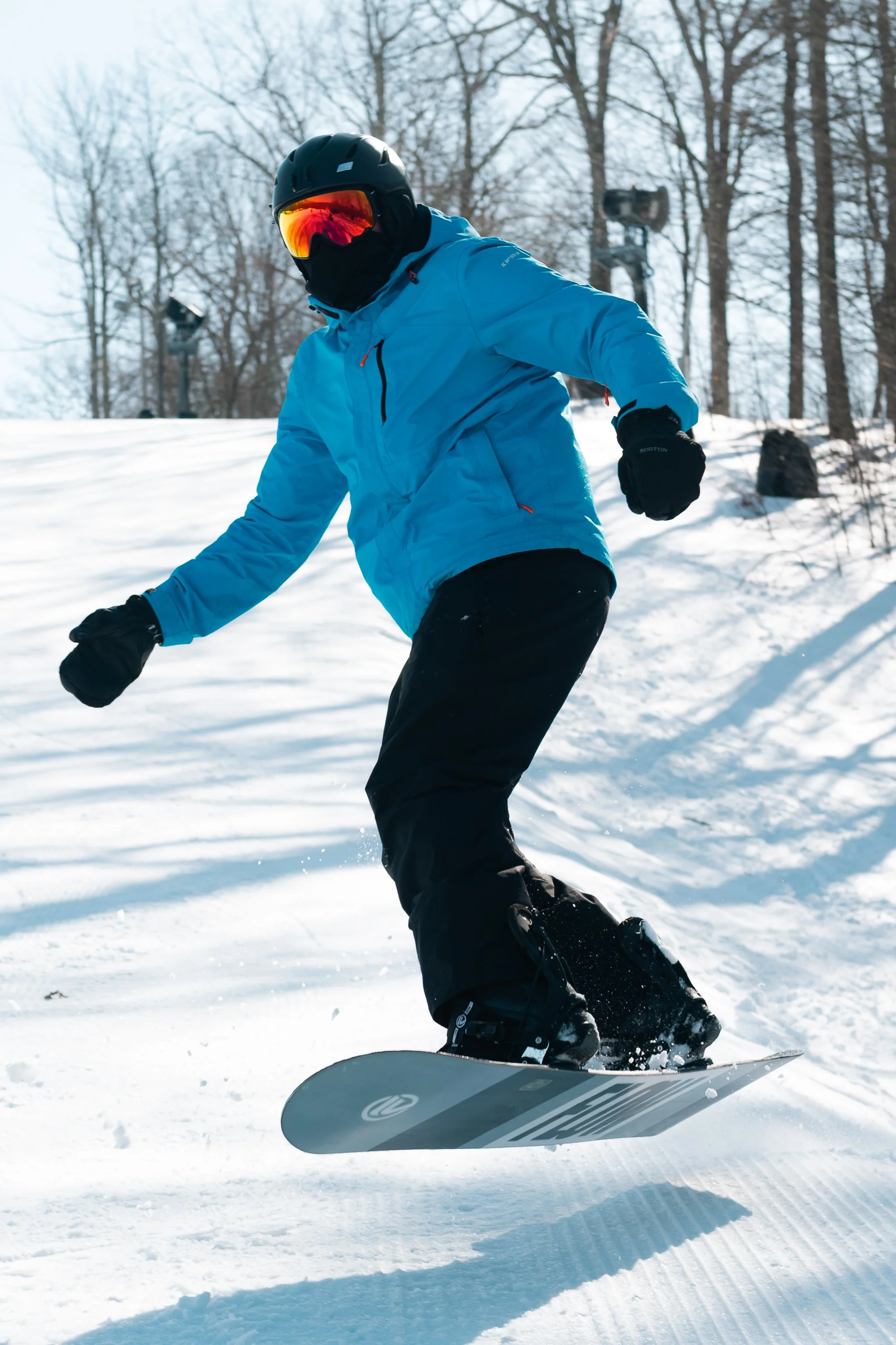 Person snowboarding on a snowy slope, wearing a blue jacket, black pants, helmet, and goggles, with trees and snow-covered ground in the background.