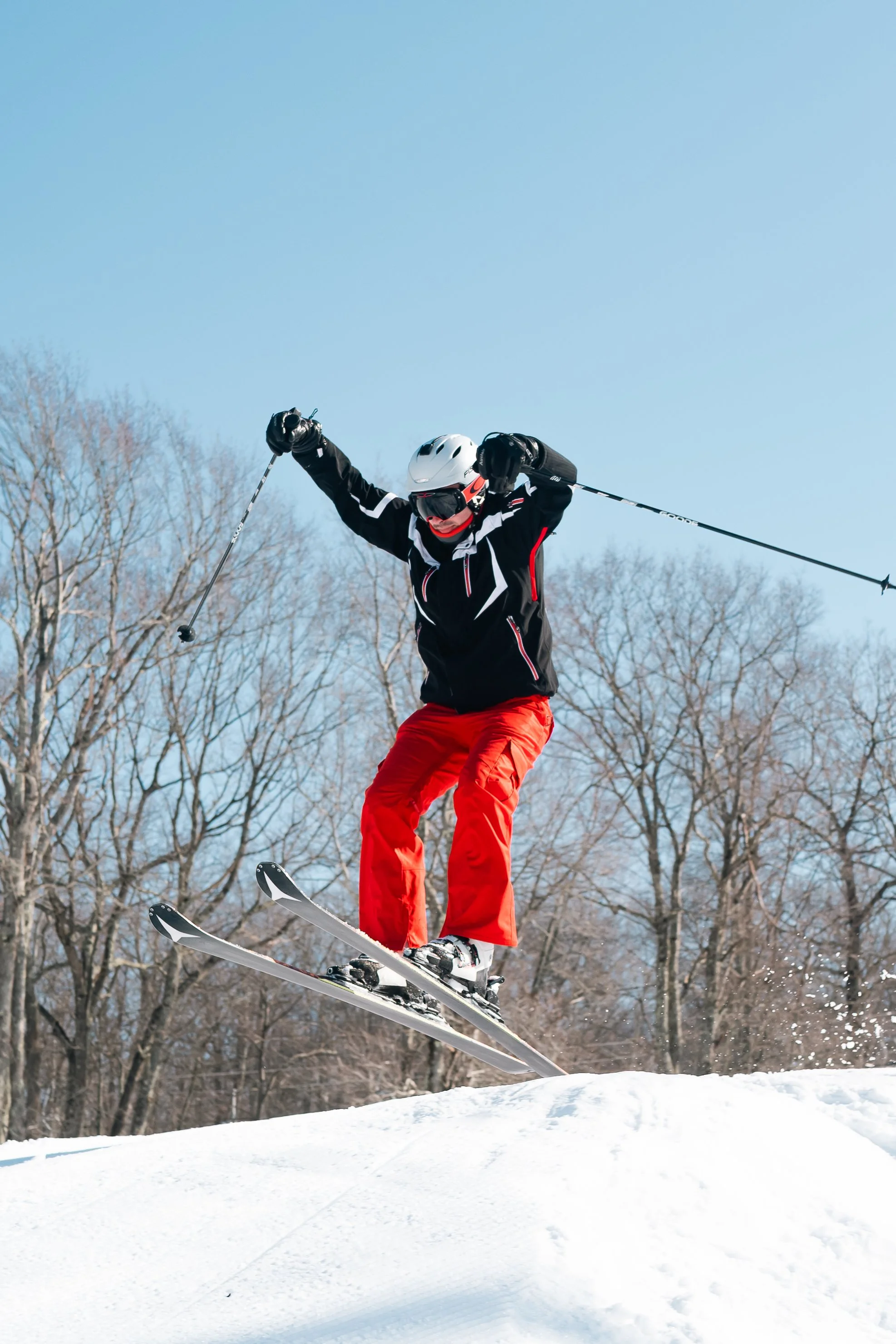 A skier in black jacket and red pants jumping off a snow ramp with bare trees and a clear blue sky in the background.