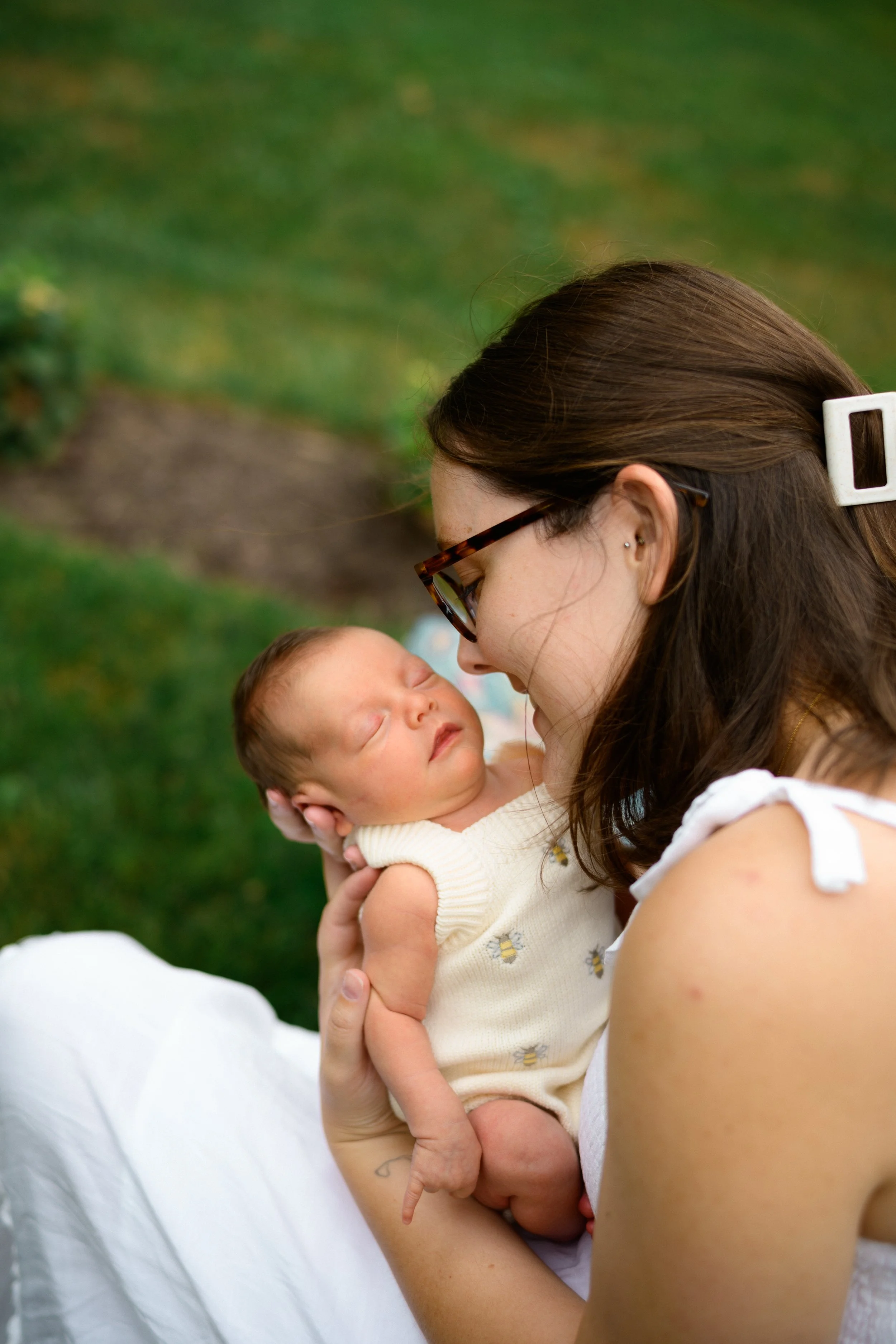 A woman with brown hair, glasses, and earrings holds a peacefully sleeping baby outdoors. The background is blurred green grass.