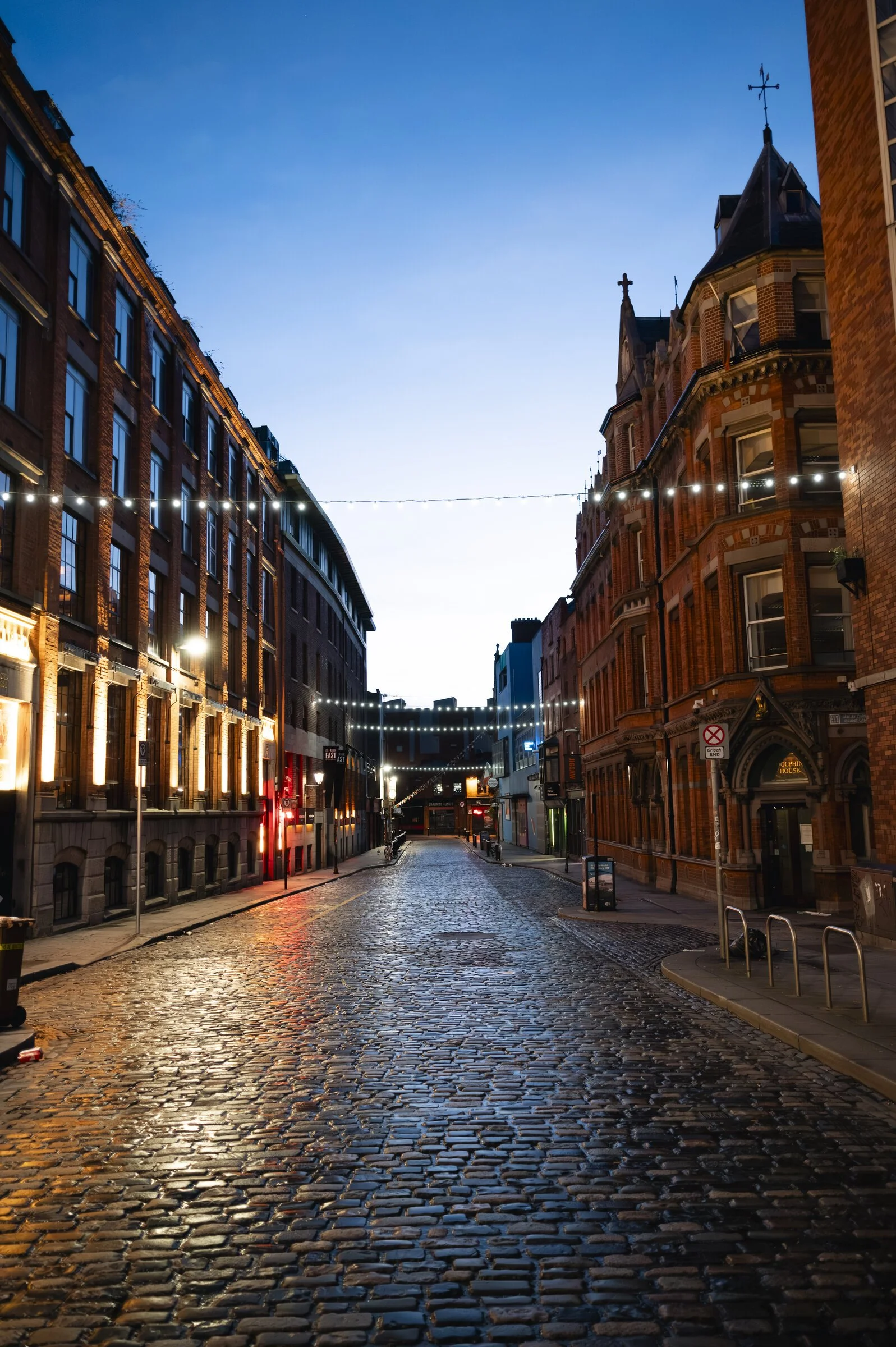 Empty cobblestone street in a city with historic brick buildings, string lights overhead, early evening with a clear sky.