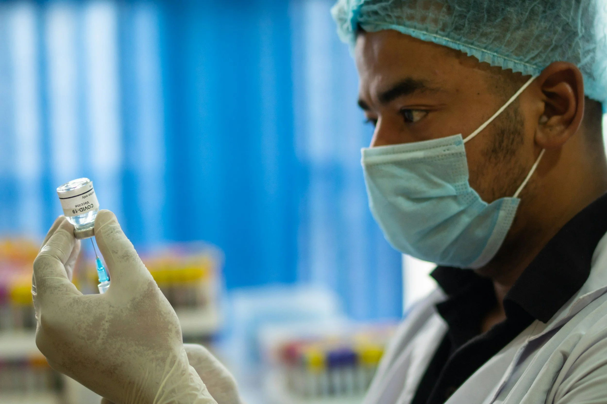 A healthcare worker wearing a mask, gloves, and a hair cap holding a vaccination vial and syringe.