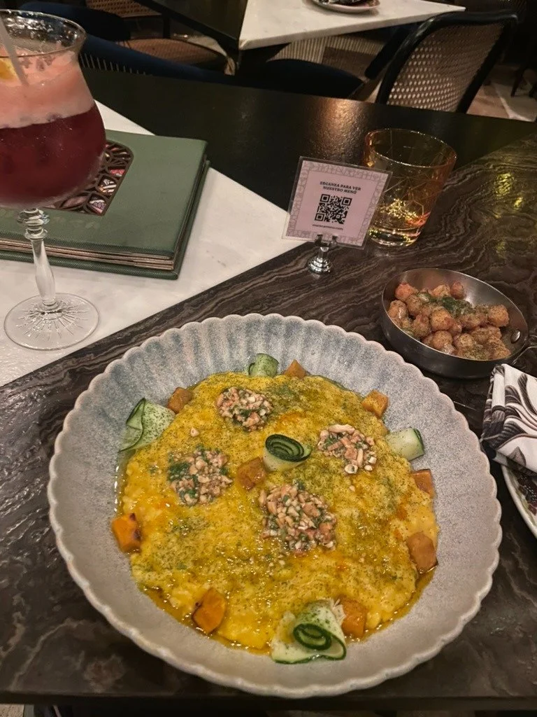 A plate of creamy scrambled eggs topped with diced vegetables, garnished with sliced cucumber and rolled cucumber slices, served at a restaurant table with a glass of red cocktail, a tumbler of whiskey, a small bowl of fried chicken, and a QR code ca