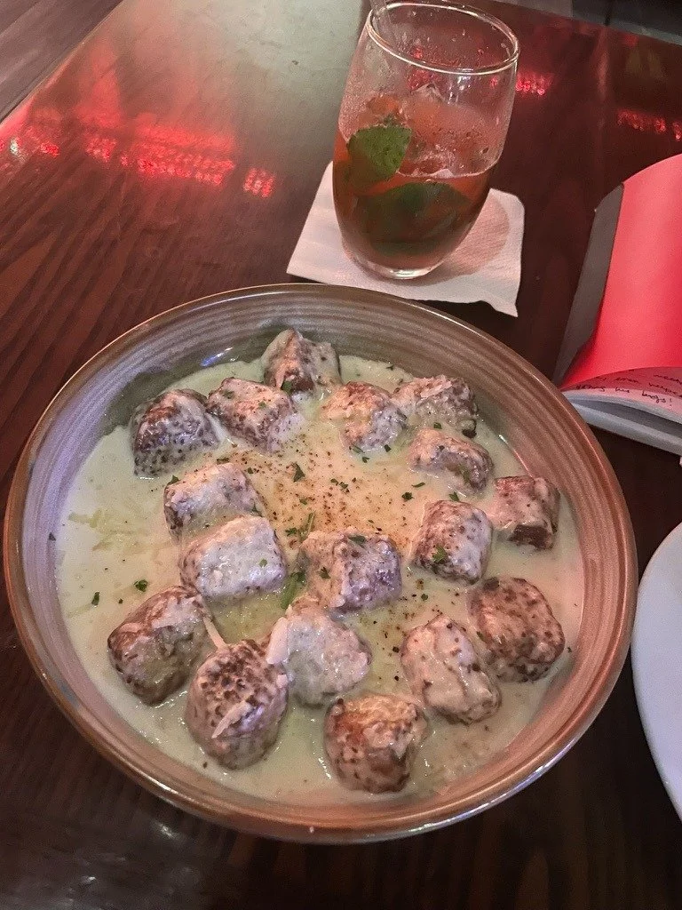 A bowl of meatballs in a creamy white sauce, garnished with herbs, on a wooden table.