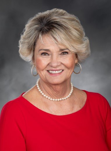 Smiling elderly woman with short blonde hair, wearing a red top, pearl necklace, and hoop earrings, against a gray background.