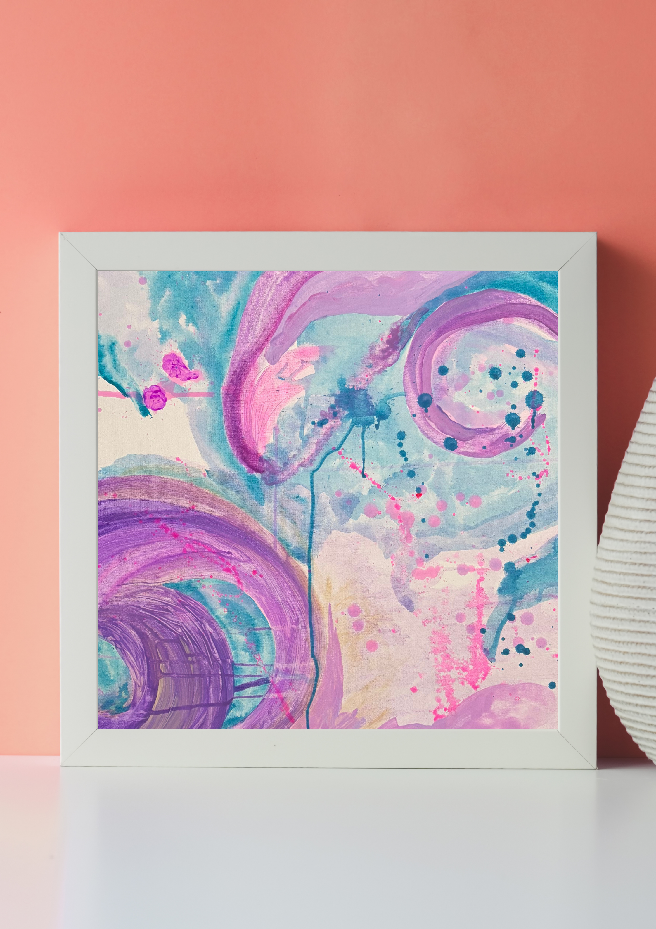 Colorful abstract painting with pink, purple, blue, and white swirls and splashes inside a white frame, against a pink wall.
