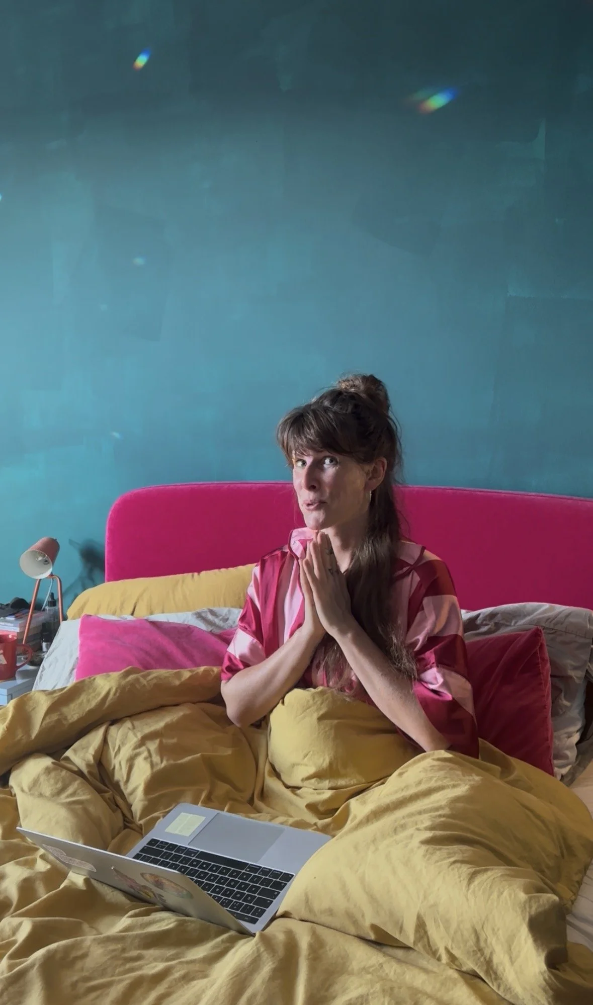 A woman sitting in bed with her hands clasped in prayer, wearing a pink and red striped top, with a yellow blanket and pink pillows, in a bedroom with teal walls, a pink headboard, a bedside lamp, a laptop, and a phone.