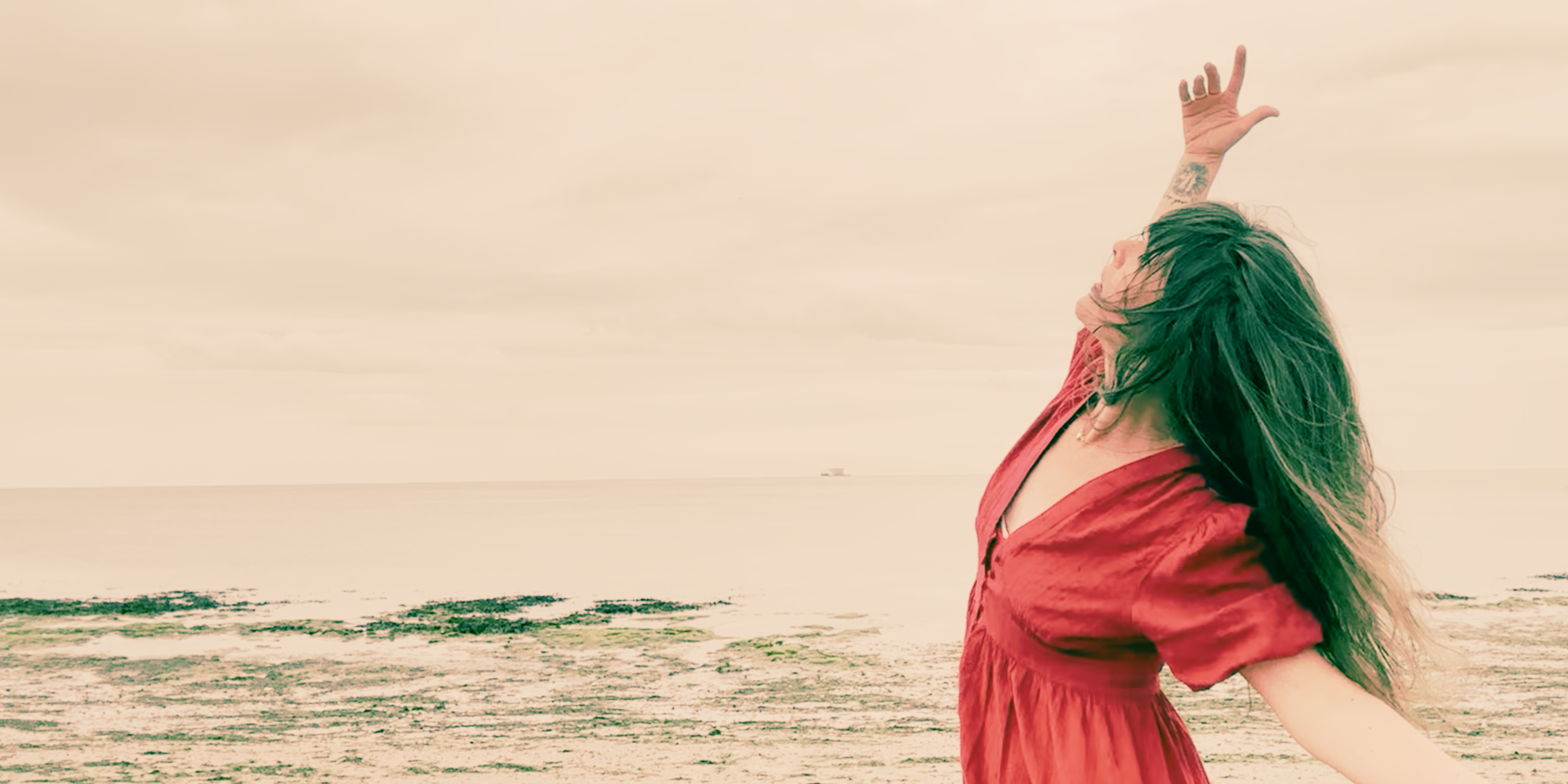 A woman with long dark hair and tattoos on her arm stands on a beach with her arm raised. She is wearing a red dress and appears to be enjoying the moment as the ocean and a cloudy sky are in the background.