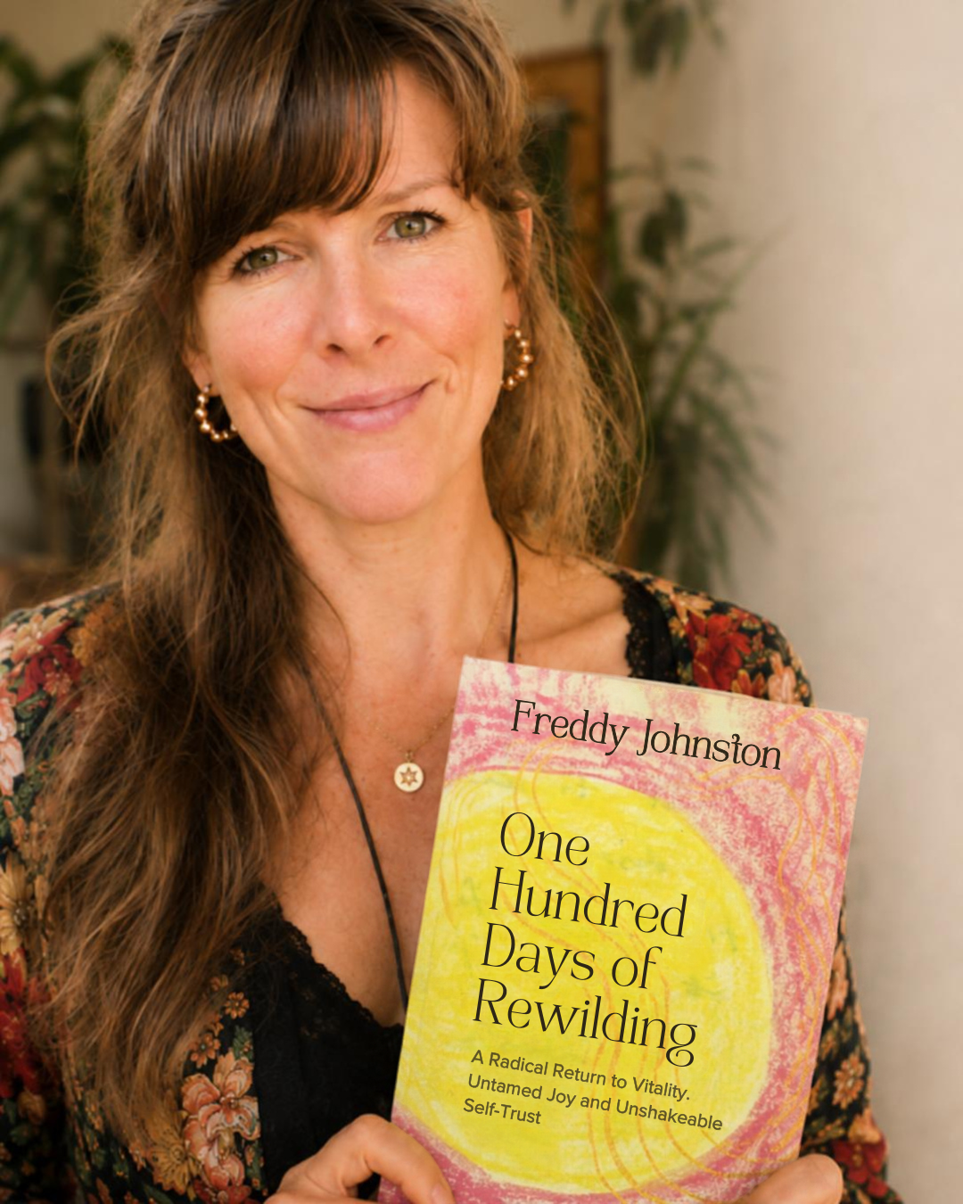 A woman with long brown hair, wearing earrings and a necklace, smiling and holding a book titled 'One Hundred Days of Rewilding' by Freddy Johnston, with a plant in the background.