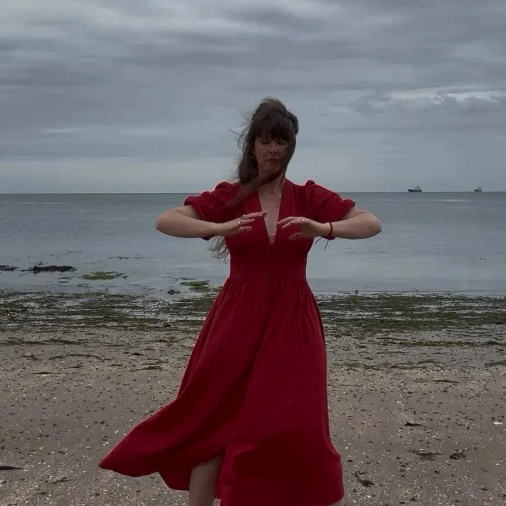 A woman in a red dress practicing a dance pose on a beach against a cloudy sky, with ships visible in the distance.