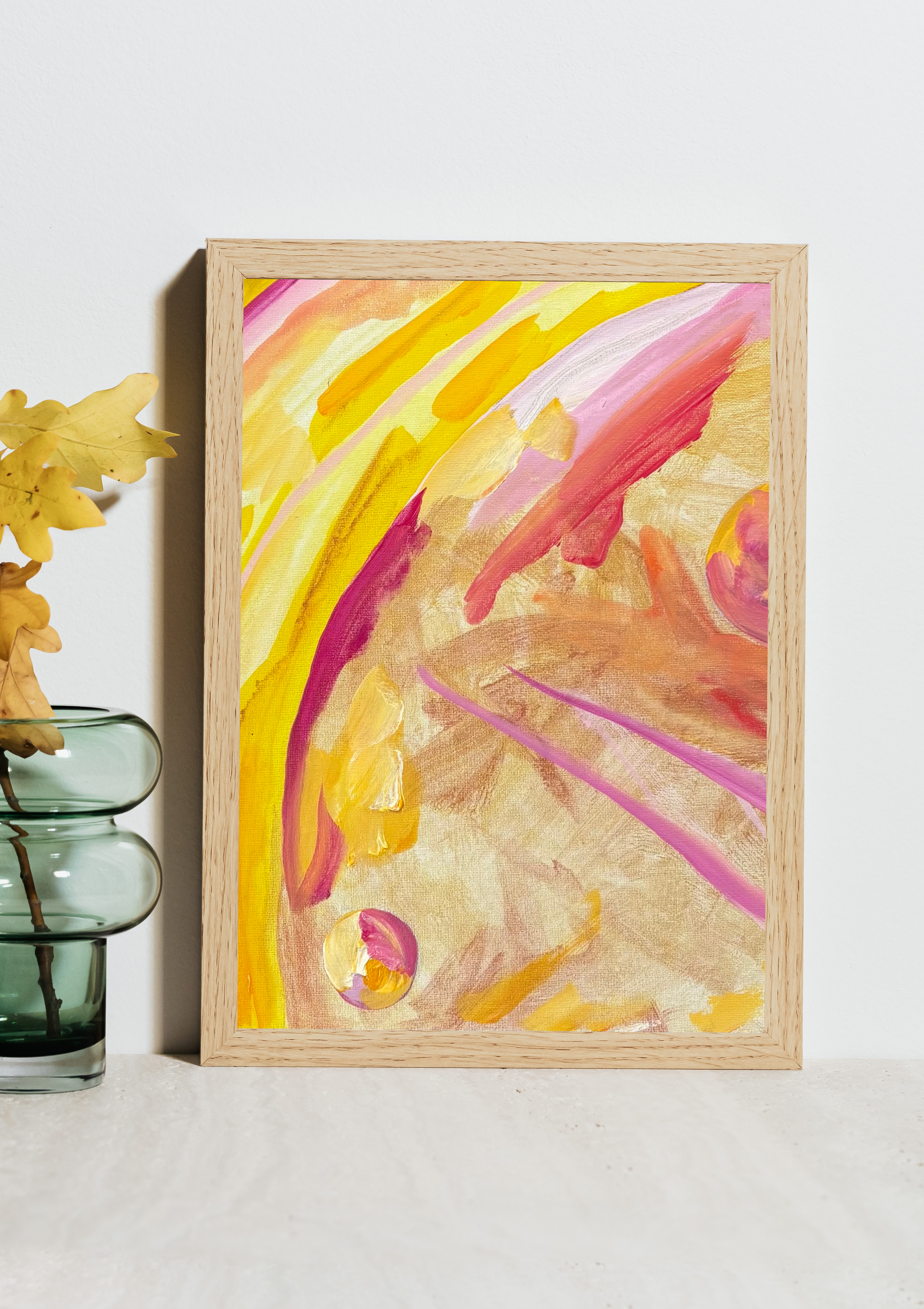 Close-up of a colorful abstract watercolor painting with pink, yellow, and orange strokes in a wooden frame, placed on a white surface with a glass vase containing yellow leaves to the left.