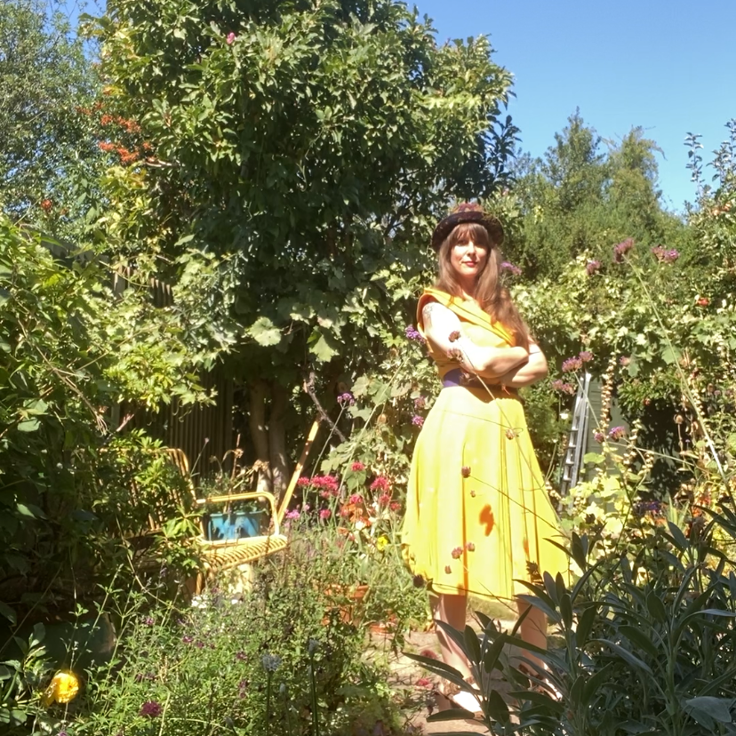 A woman standing in a lush garden surrounded by various plants and flowers, wearing a yellow skirt, white top, and a hat under a clear blue sky.