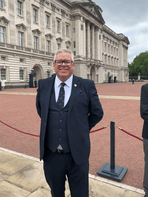 A smiling man in a navy suit with glasses and a patterned tie, standing outdoors in front of a large historic building with ornate architecture.