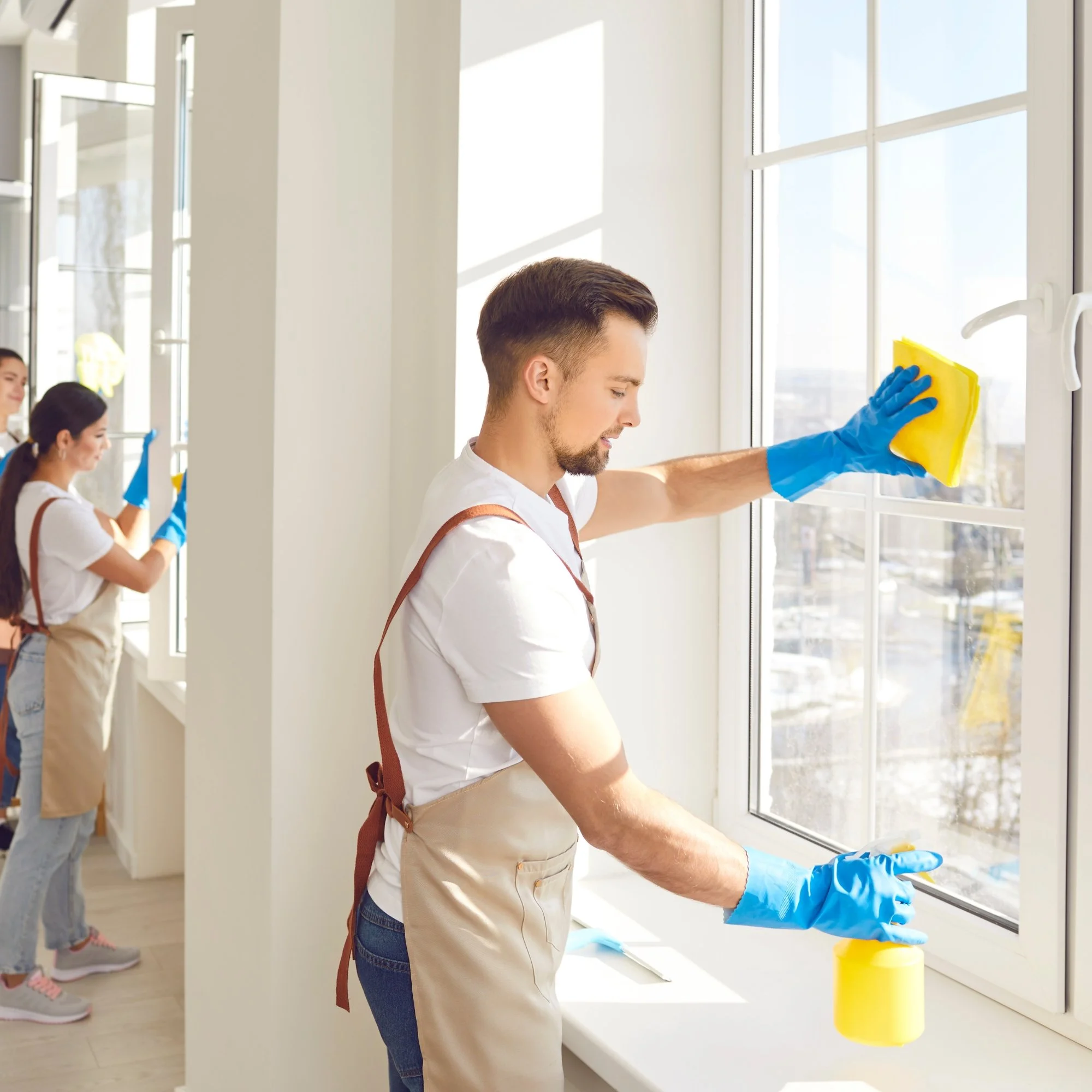 A man wearing blue gloves, a white t-shirt, and an apron cleaning a window with a yellow spray bottle and yellow sponge. Two women in the background wearing similar attire are also cleaning windows.