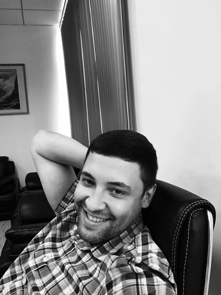 A young man with short dark hair and a beard, smiling, sitting in a black leather chair with white stitching, in an office or waiting room, wearing a checkered shirt, with a relaxed pose and his arm behind his head.