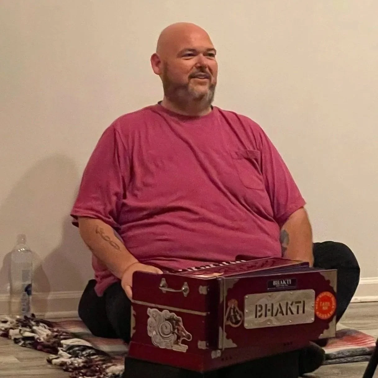 A man with a bald head and beard, wearing a red t-shirt, sitting on a rug on the floor next to a cardboard box, with a water bottle nearby, against a plain wall.