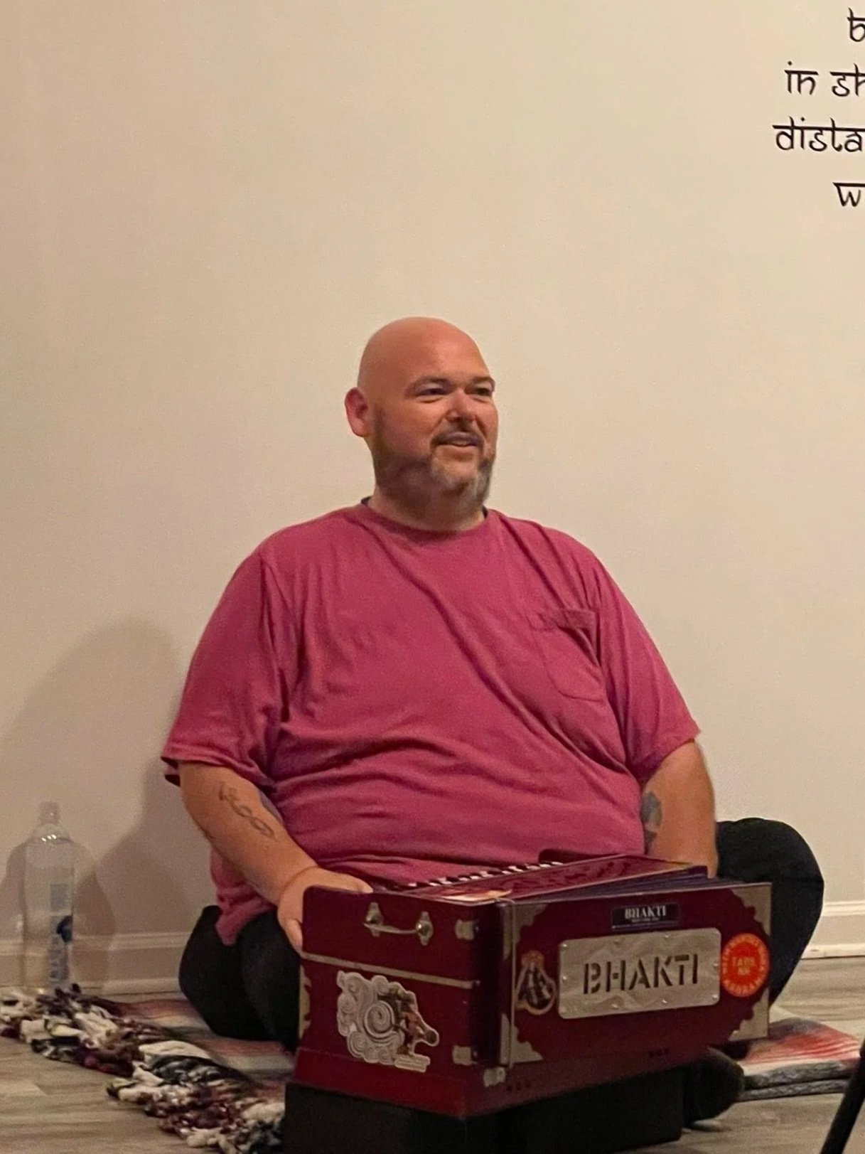 A man with a beard and shaved head, wearing a red t-shirt, sitting on the floor and playing a harmonium labeled 'Bhakti' with a pink and white patterned cloth underneath. A water bottle is visible in the background to the left.
