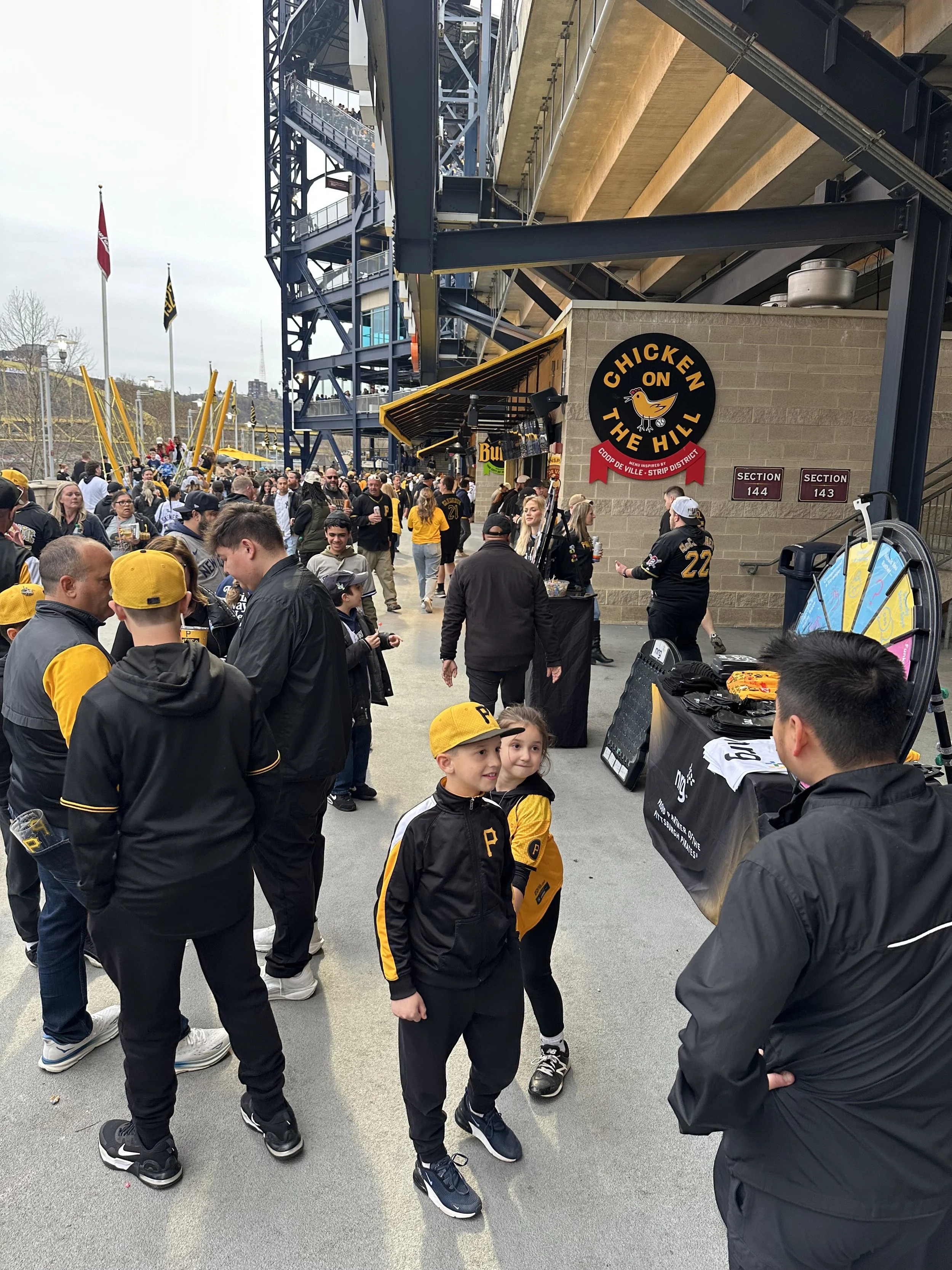 Outside of a stadium with fans in baseball-themed apparel, including children wearing Pittsburgh Pirates hats, gathered for an event near the entrance, with signs for food and a prize wheel in the background.