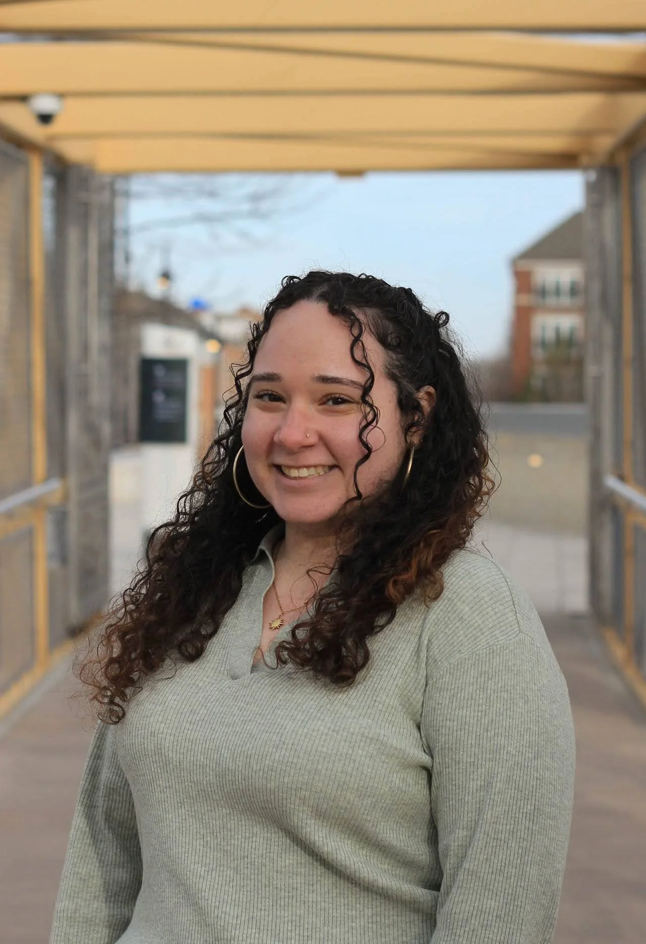 A smiling woman with curly dark hair, large hoop earrings, a nose piercing, wearing a light-colored ribbed sweater, standing under a yellow wooden structure outdoors during daytime.