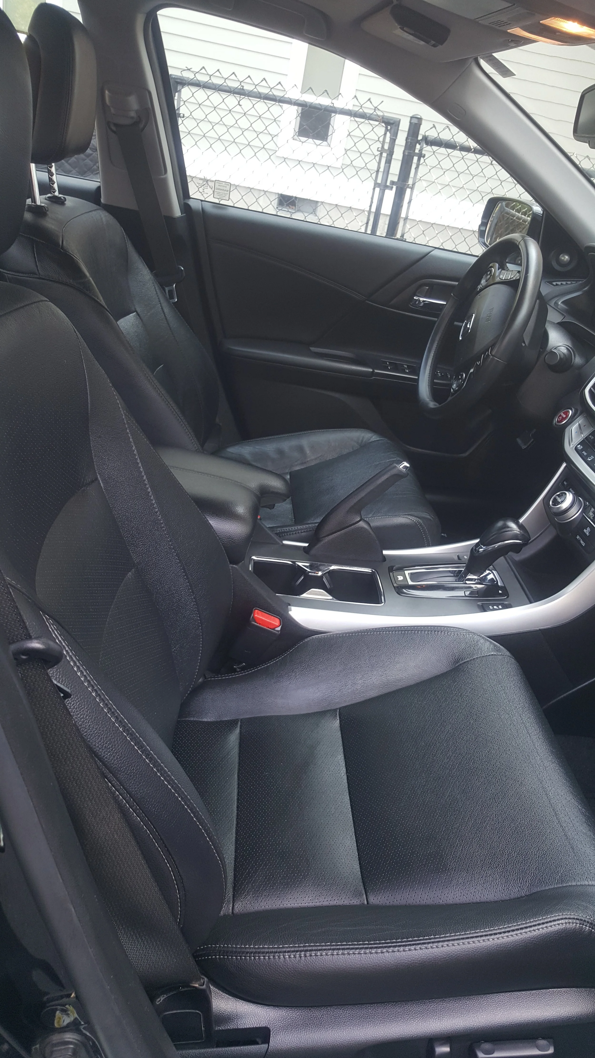 Interior of a car with black leather seats, center console, steering wheel, and dashboard, parked behind a chain-link fence near a house.