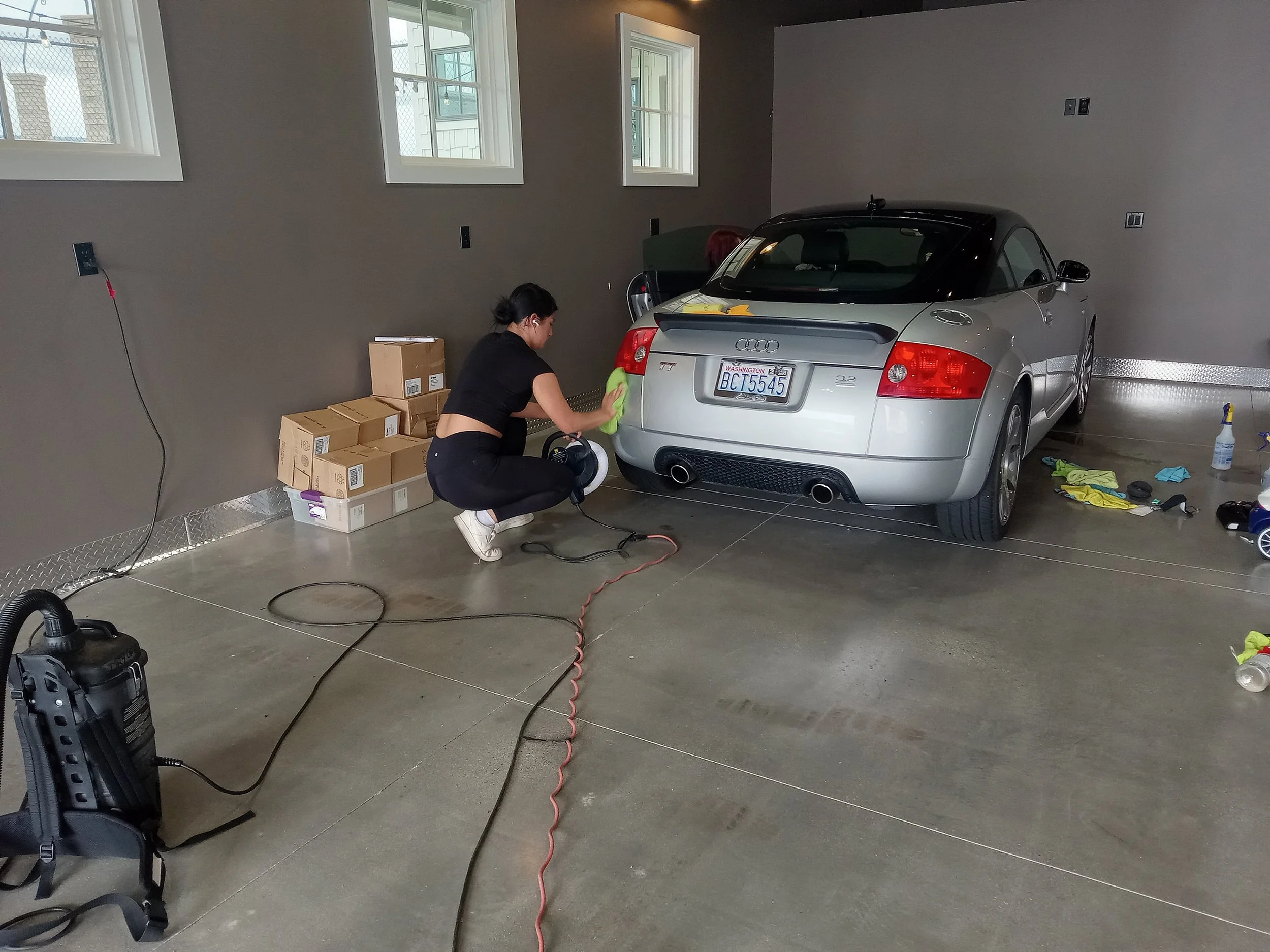 Woman cleaning a silver convertible car inside a garage, with cleaning supplies and boxes nearby.