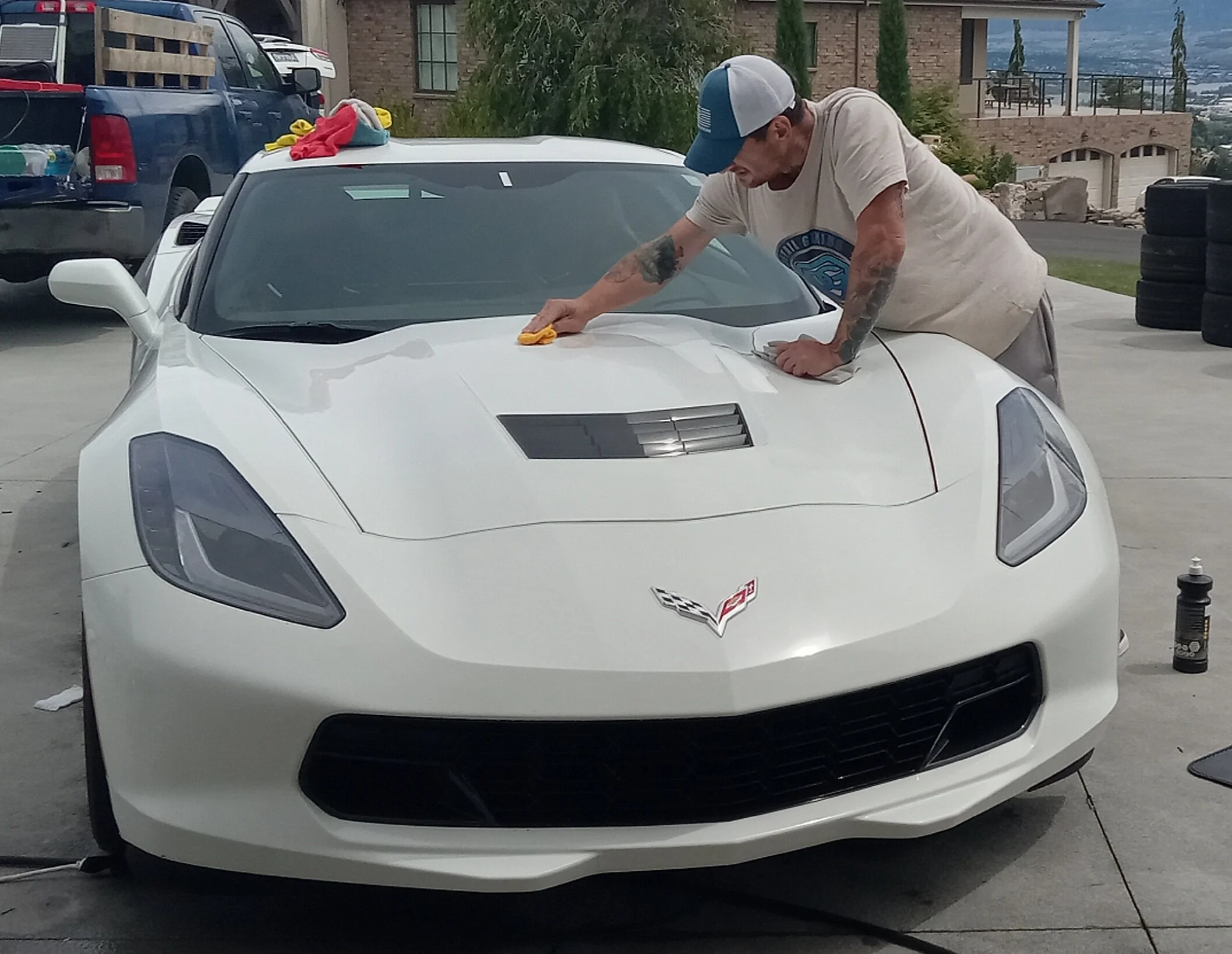 Man in beige t-shirt and baseball cap polishing a white Chevrolet Corvette with a yellow cloth in an outdoor driveway area.