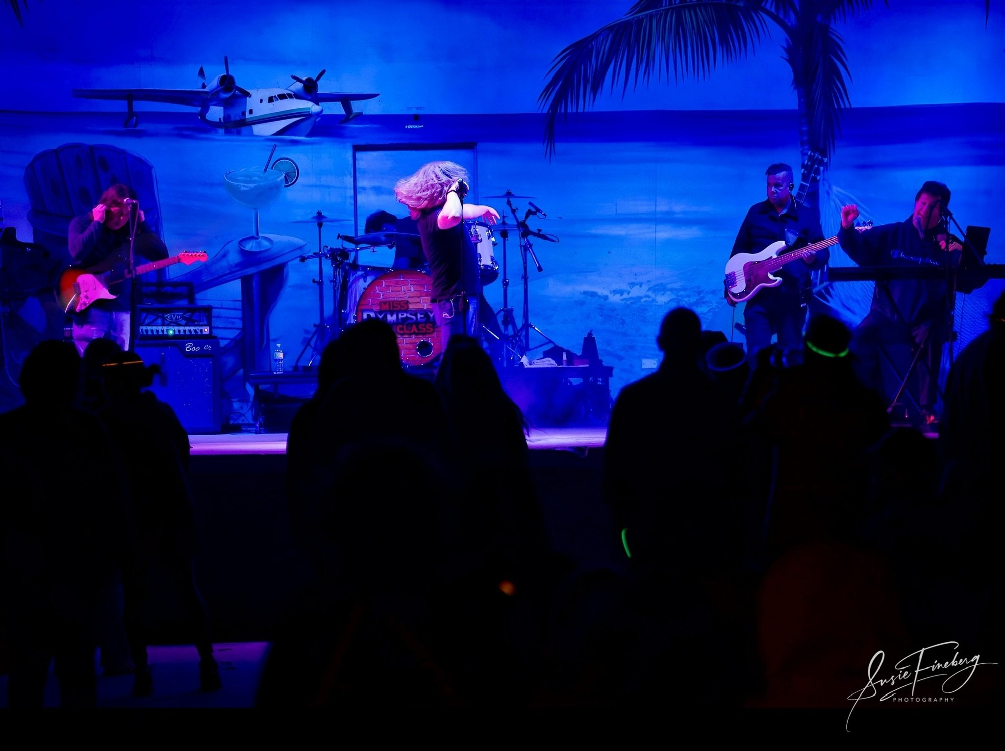 A band performing on stage with a beach-themed backdrop, including a palm tree, airplane, cocktail, and ocean scene, under blue and purple lighting.