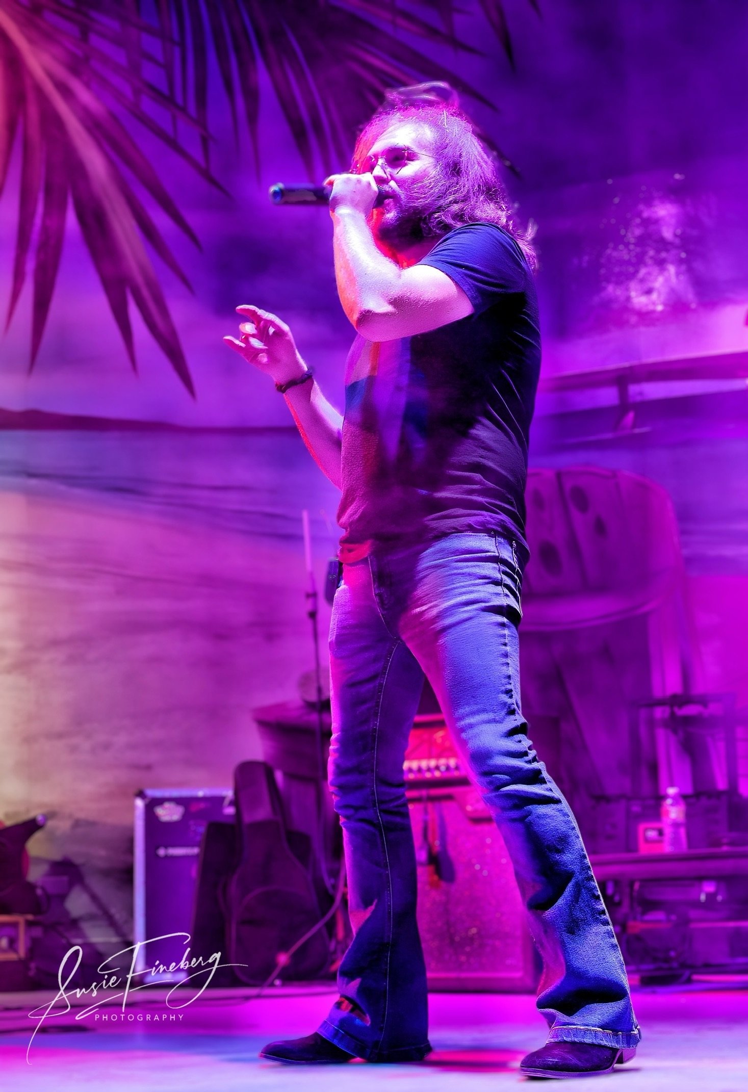 A man with long hair and beard performing on stage, holding a microphone, surrounded by purple and pink stage lighting, with a decorated backdrop and musical equipment in the background.