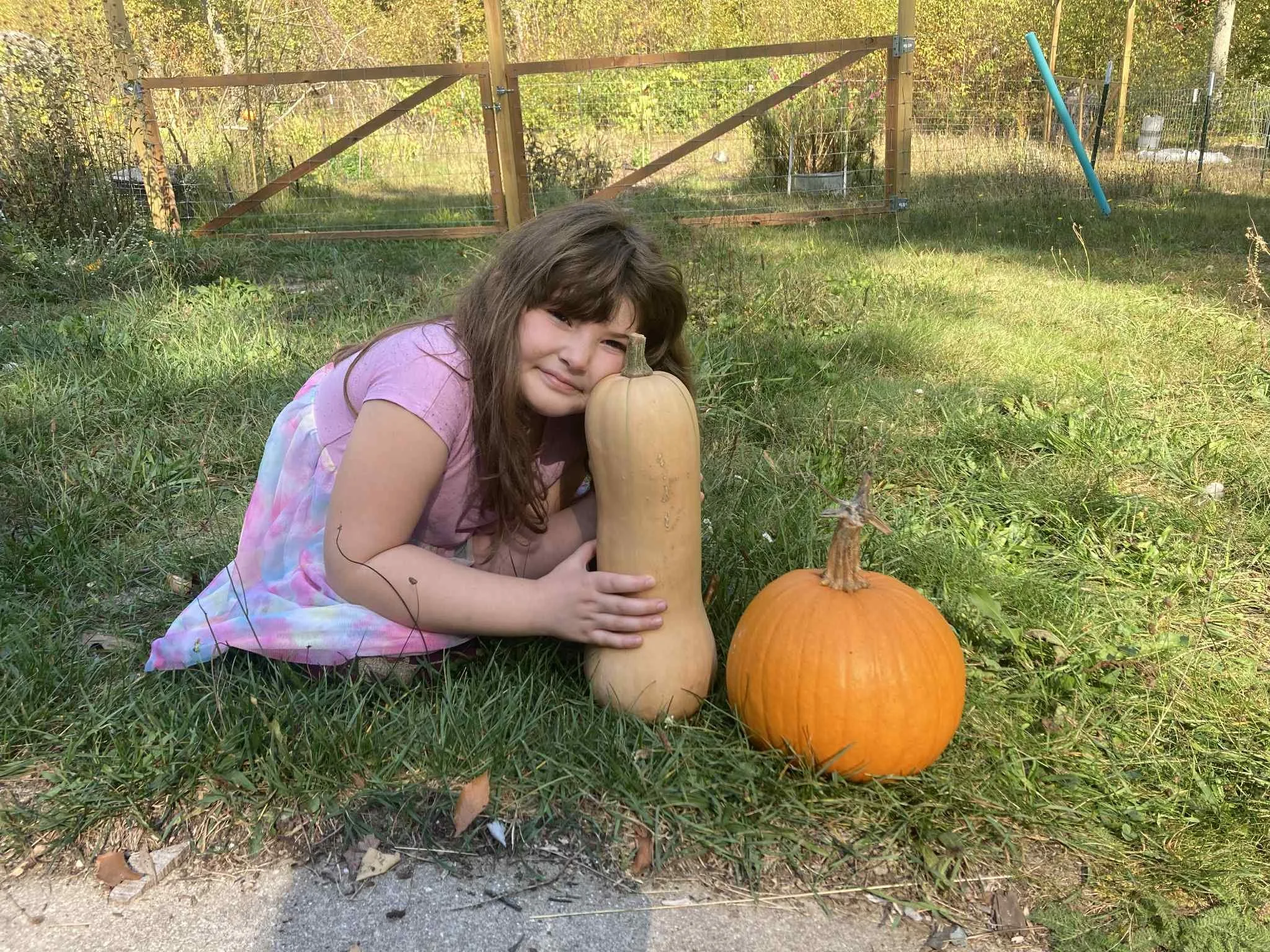 A young girl with long brown hair wearing a pink dress, lying on the grass outside, hugging a large butternut squash next to a pumpkin.