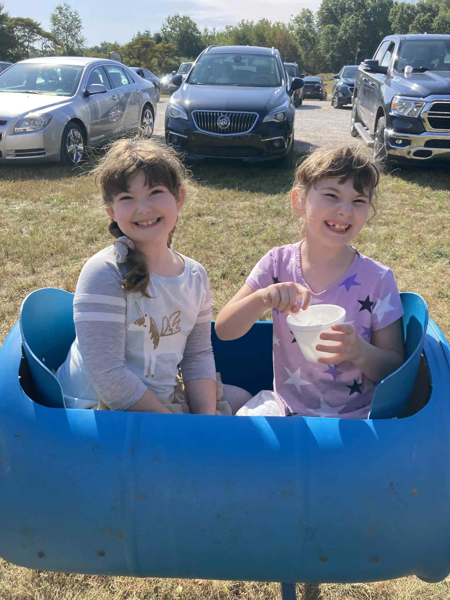 Two young girls with brown hair, sitting in a blue wagon outdoors on a sunny day, smiling at the camera. One girl is holding a cup.