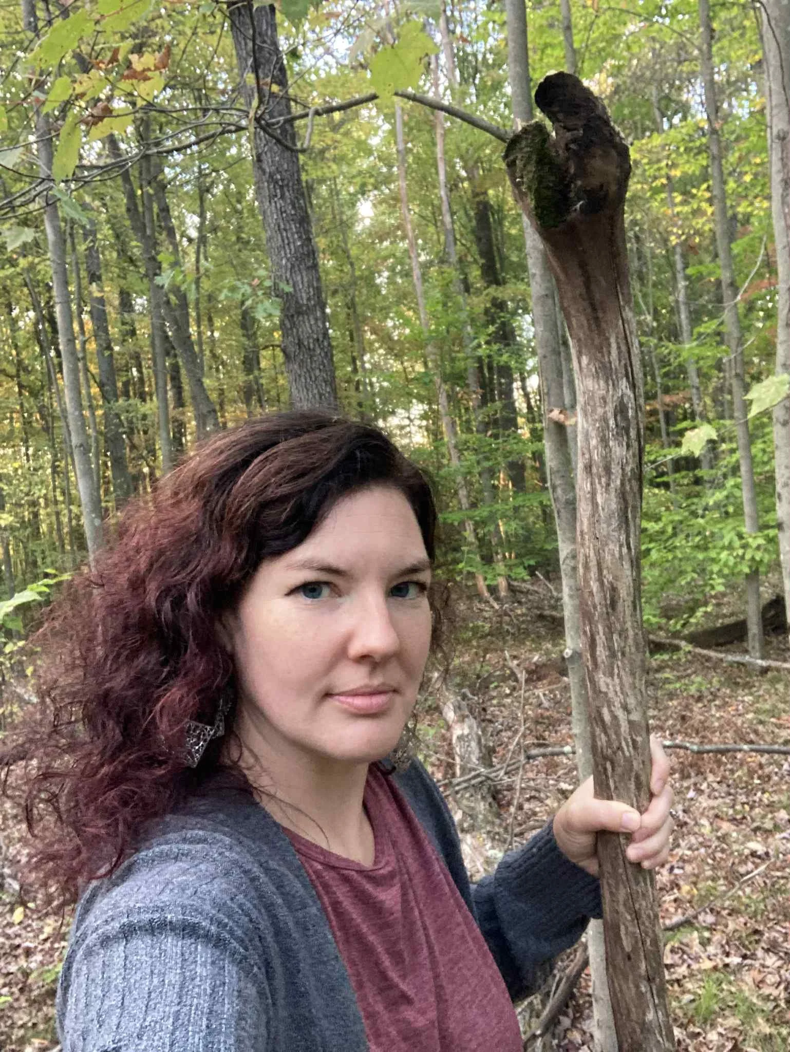 A woman with curly dark hair and light skin standing in a forest, holding a tall, split tree branch. The forest has green leaves and trees in the background.