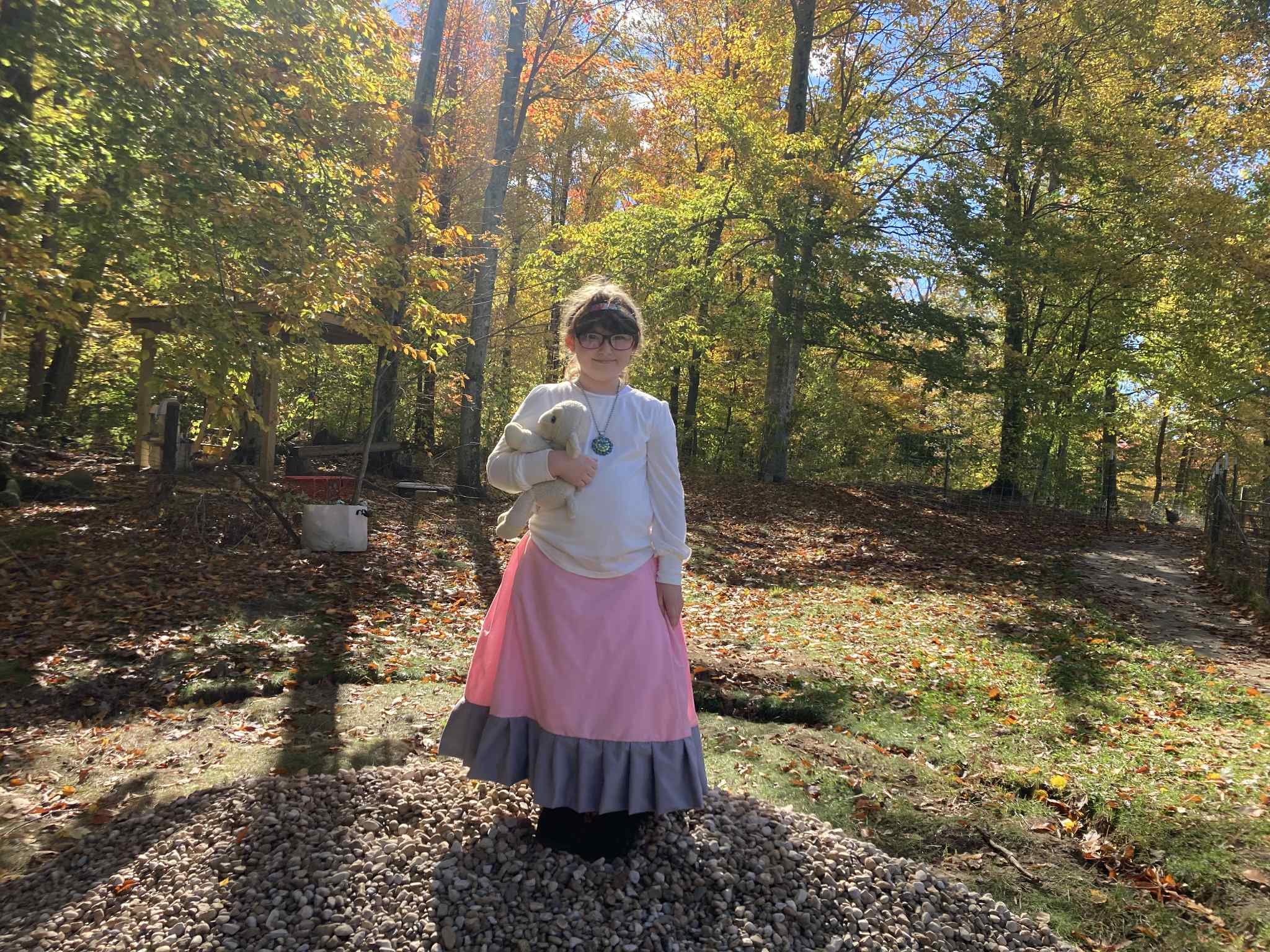 A young girl dressed in a long pink and gray skirt, white blouse, and headband, holding a stuffed animal, standing on a mound of rocks in a woodland area with colorful autumn foliage.