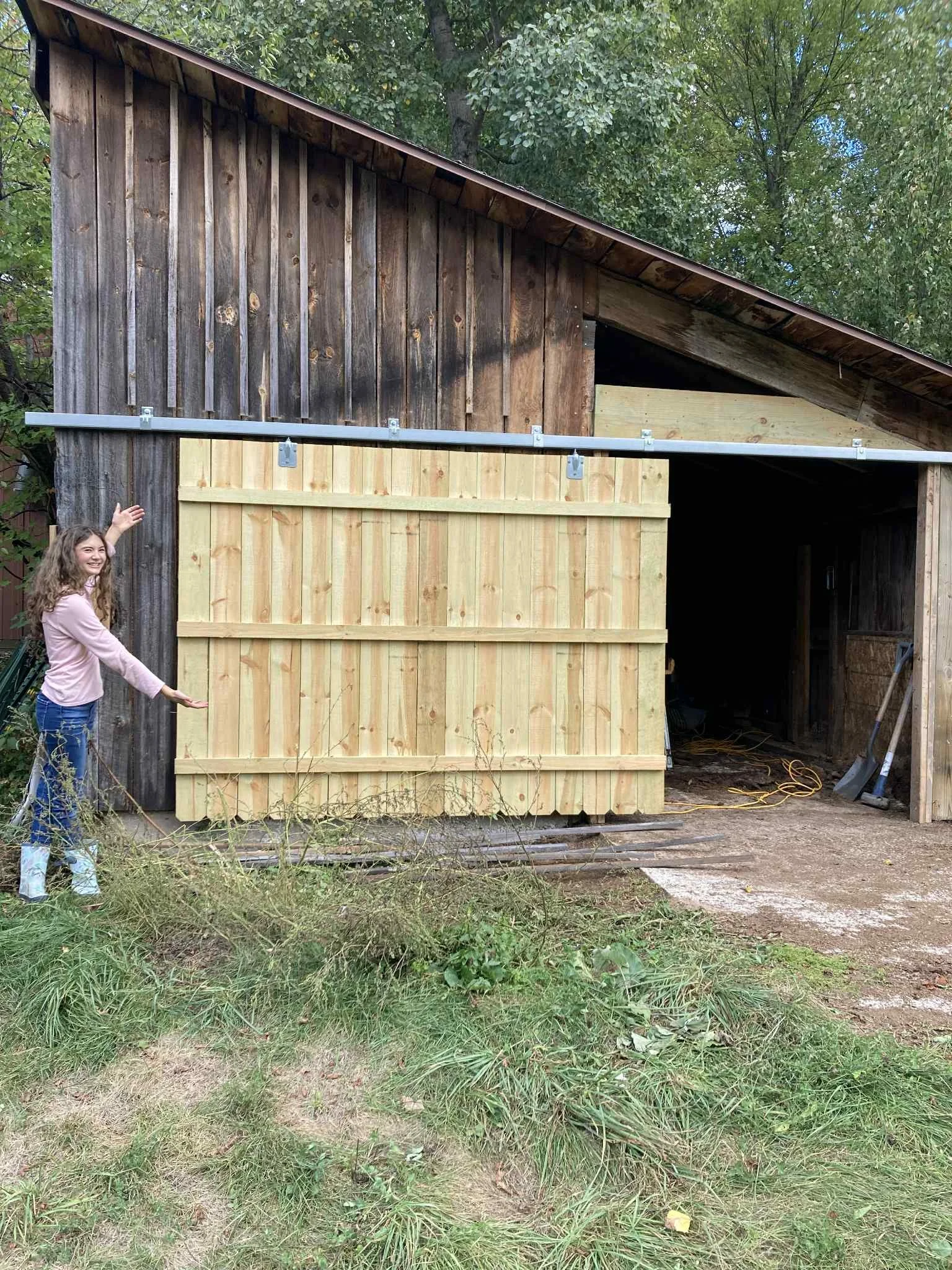 A young girl wearing a pink jacket, blue jeans, and metallic boots standing outside a wooden barn under construction, pointing towards the barn with a smile.