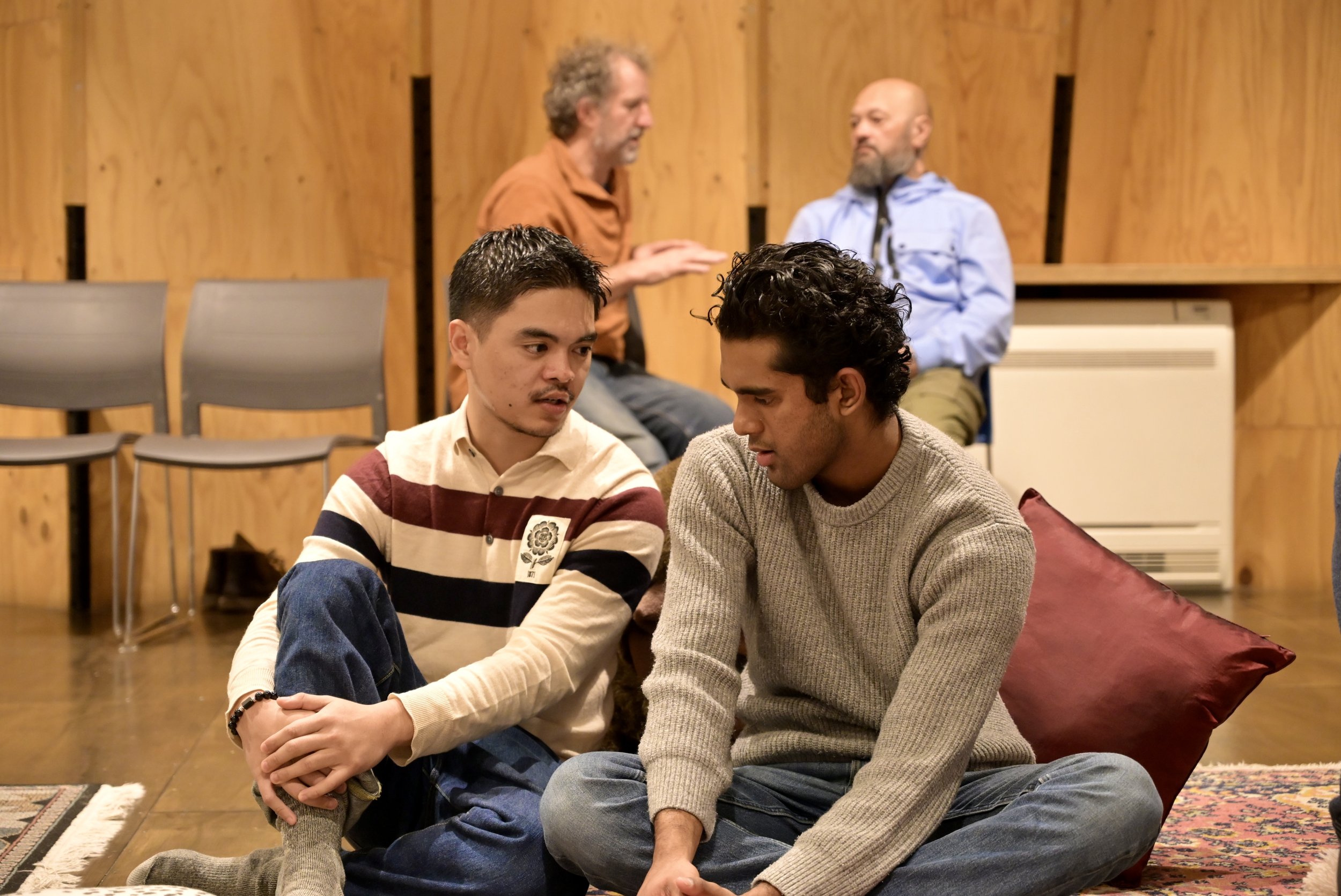 Two men sit on a carpeted floor, engaged in an honest, open hearted conversation, while two others sit apart in the background, having a separate discussion.