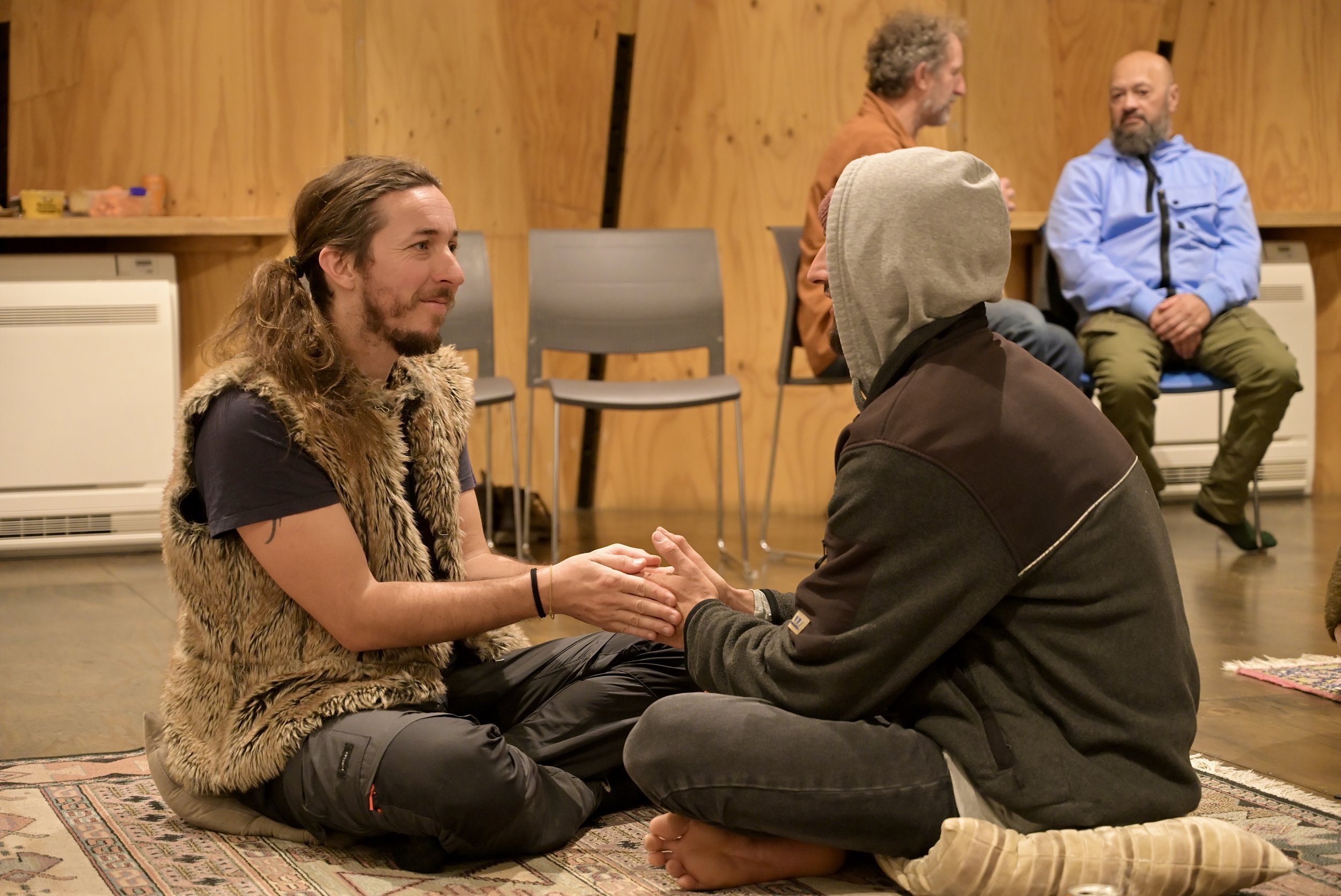 Two men sit on a rug, holding hands and facing each other, in a room with wooden walls. One man has long hair, a beard, and wears a faux fur vest. The other man wears a hoodie pulled over his head. Three other men are sitting in the background, obser