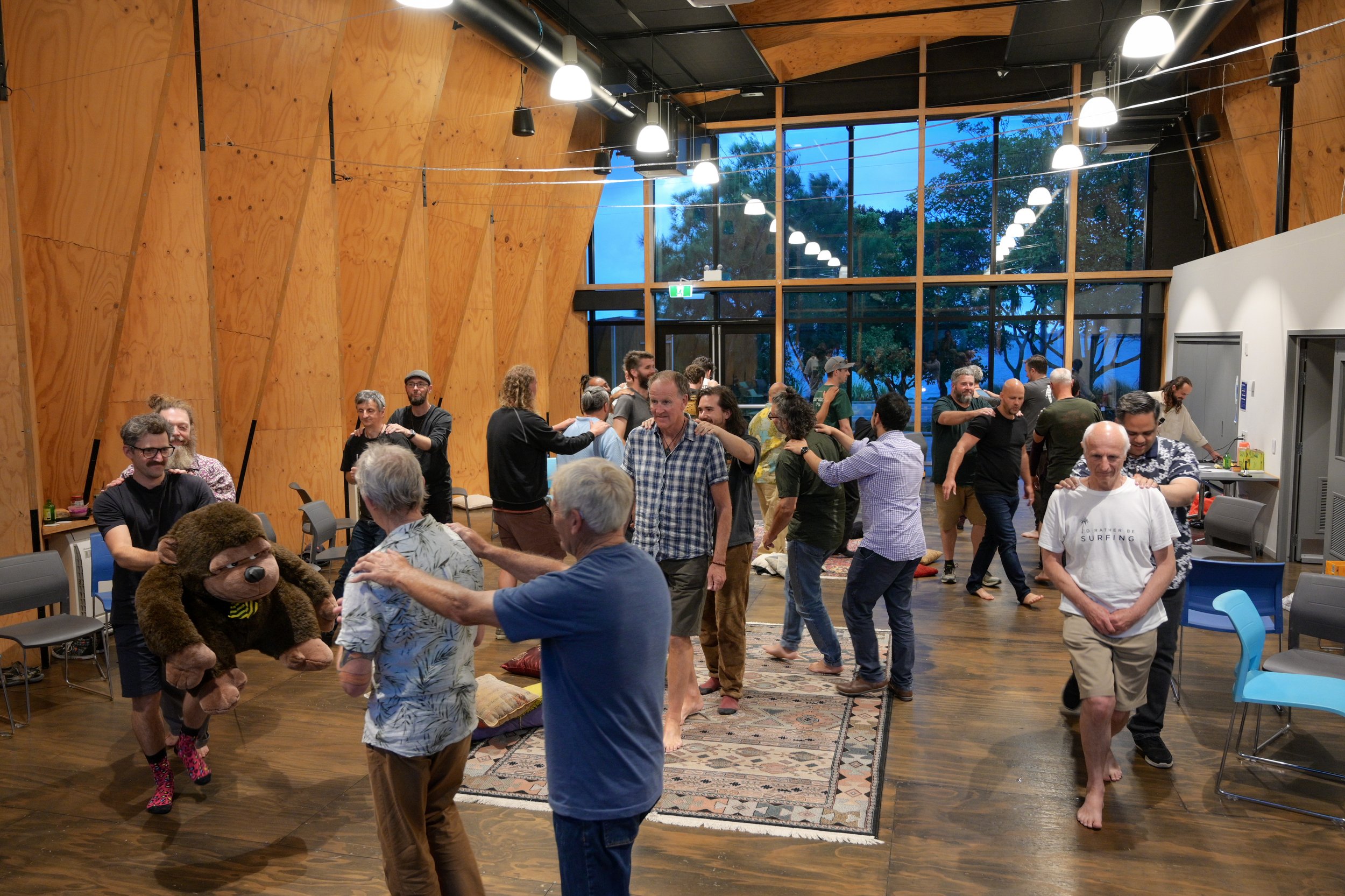 People dancing and socializing in a large, warmly lit, wooden-walled room with high ceilings and large glass windows at dusk.