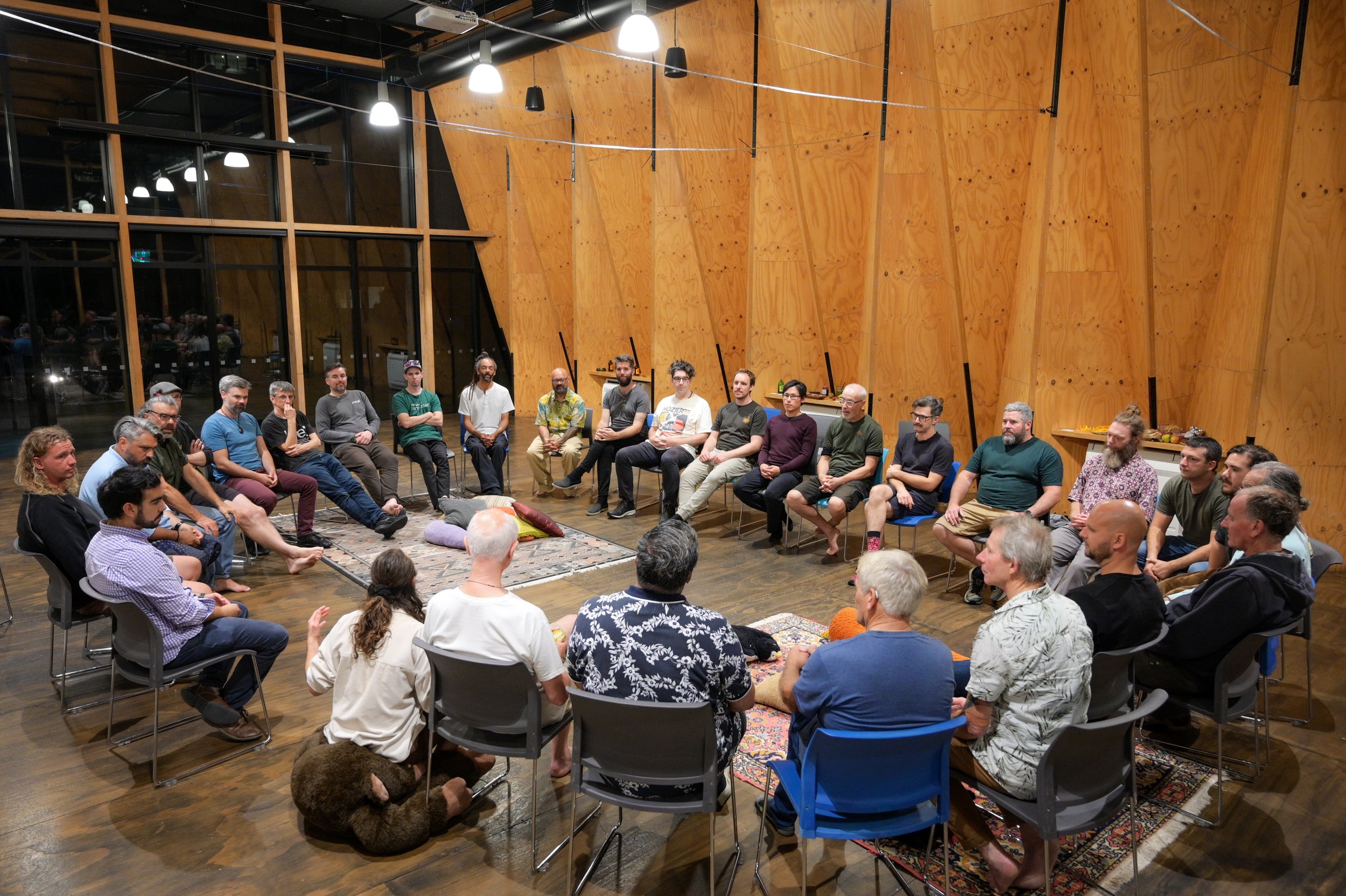 A large group of men intentionally meeting for men's work sitting in a circle. It's in a spacious, modern room with wooden walls and large windows.