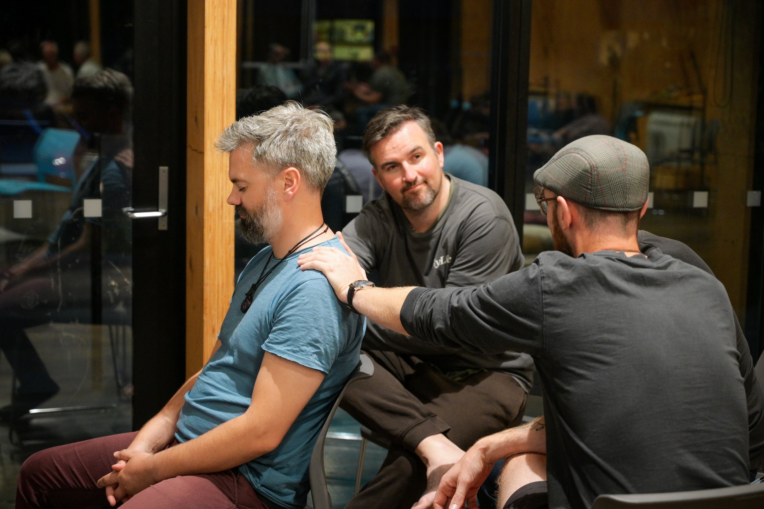 Three men sitting in a circle participate in an activity, with the man in the middle placing his hand on the shoulder of the man on the left, who has his eyes closed.